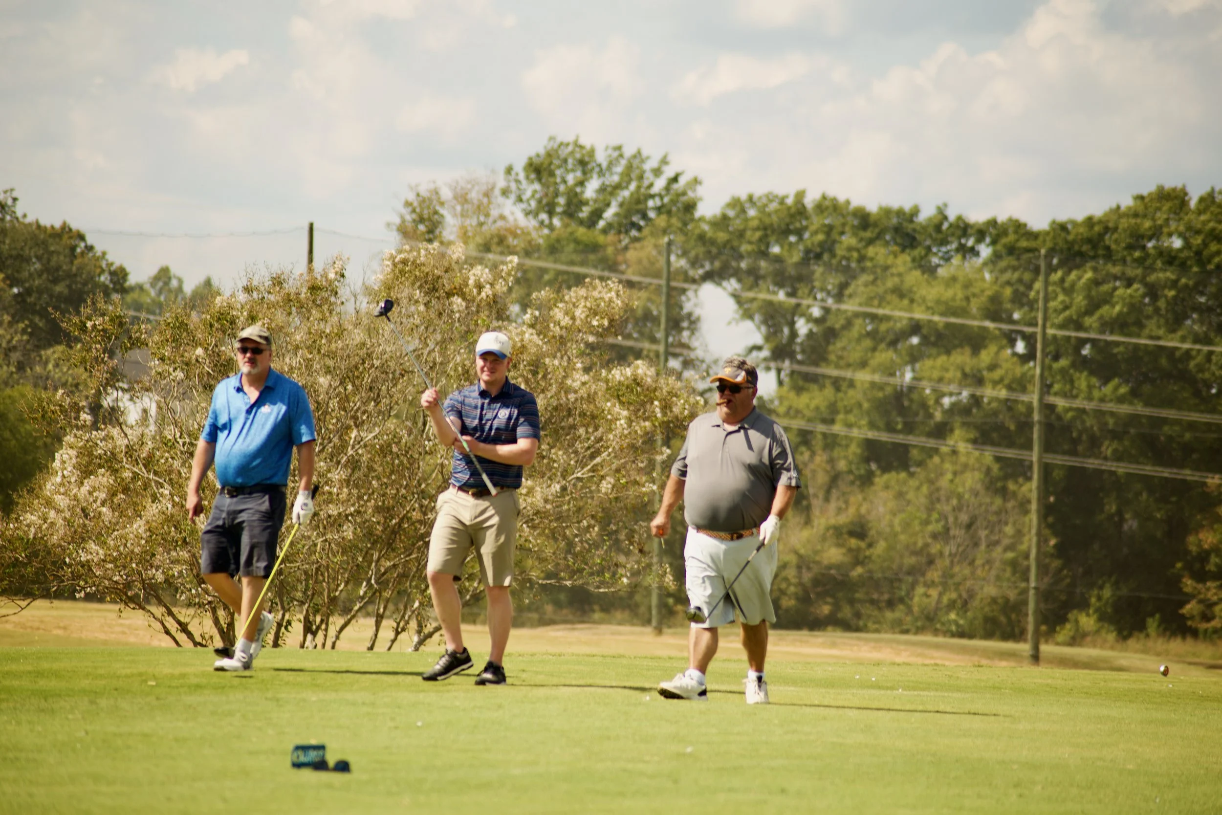 Four men on a golf course, walking and preparing to putt a golf ball. They are wearing casual golf attire and hats, with trees and power lines in the background.