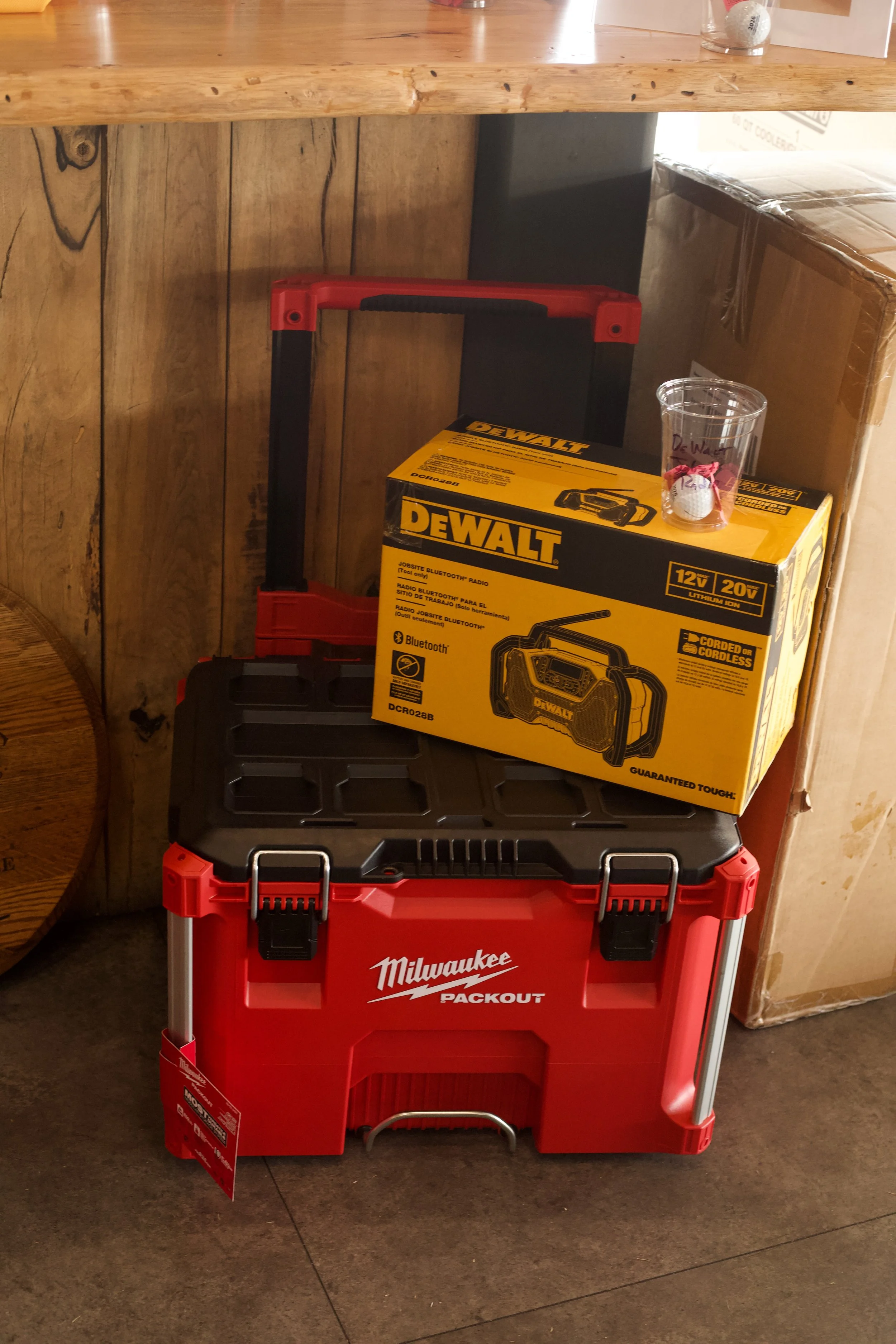 Red Milwaukee Packout toolbox on a concrete floor, with a yellow Dewalt box, a cardboard box, a clear plastic cup with a golf ball and golf tee, and a cardboard box in the background.