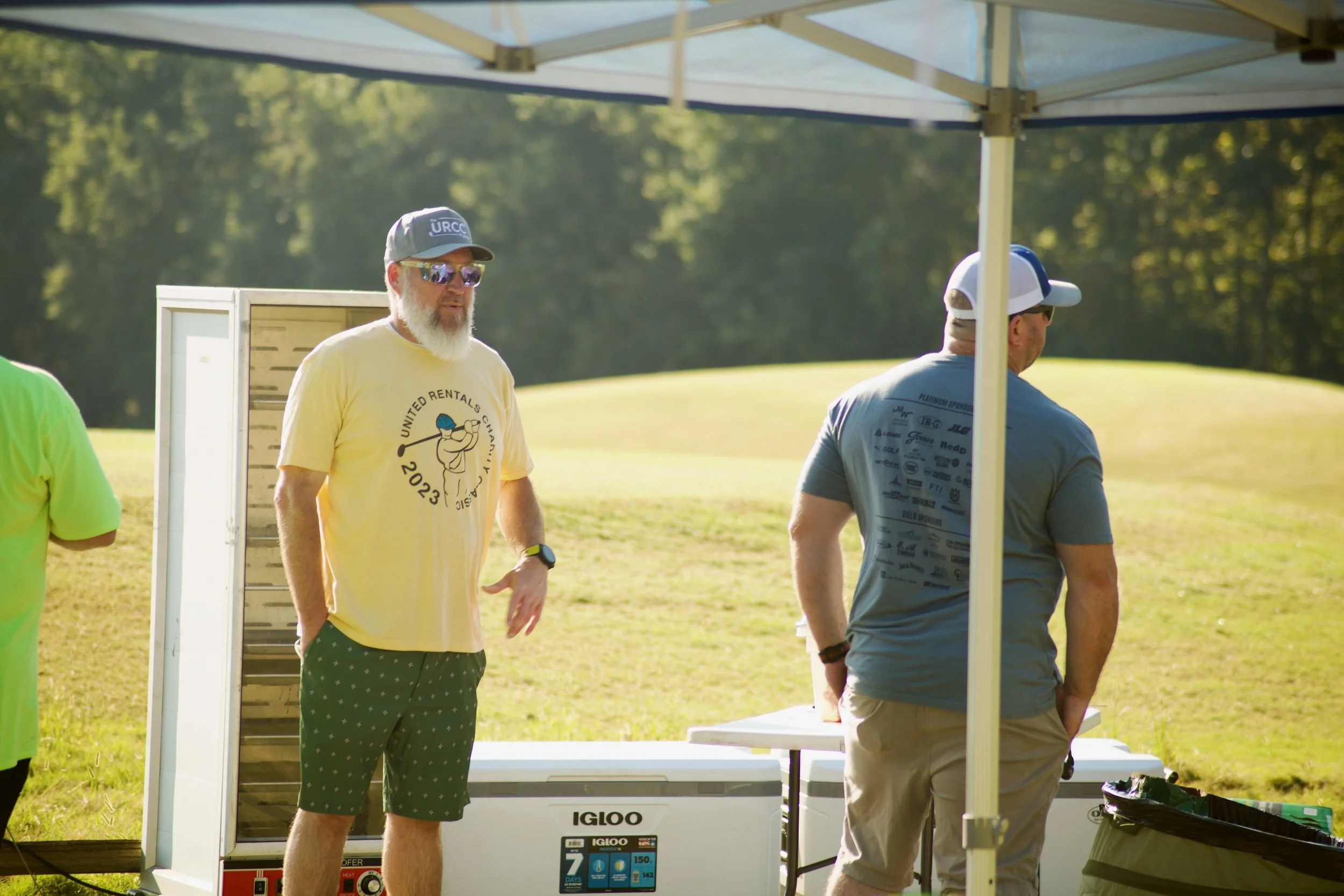 People standing outdoors under a canopy in a park, with a portable generator and trees in the background.