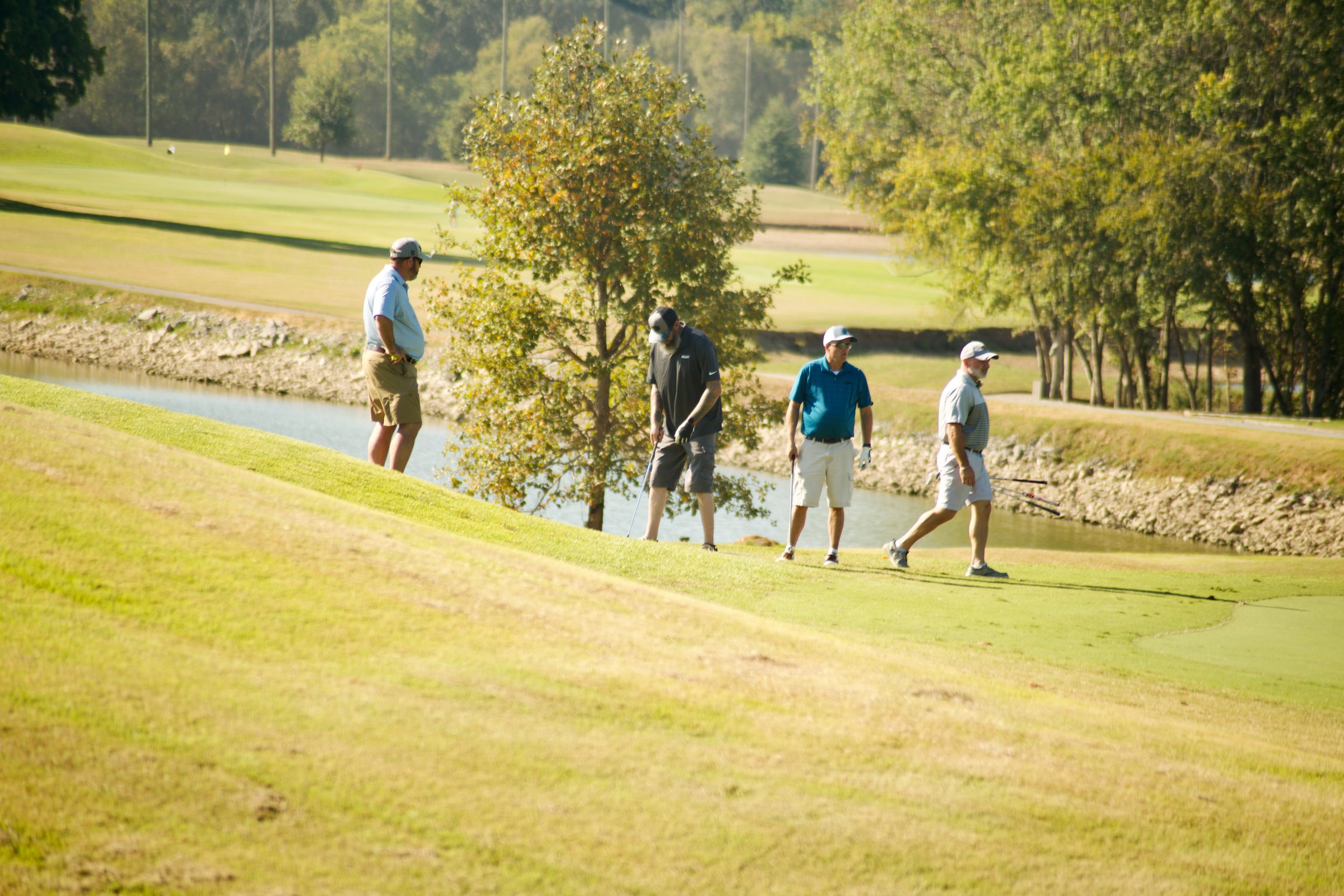 Four men walking along a golf course near a small waterway, surrounded by trees and grassy areas on a sunny day.