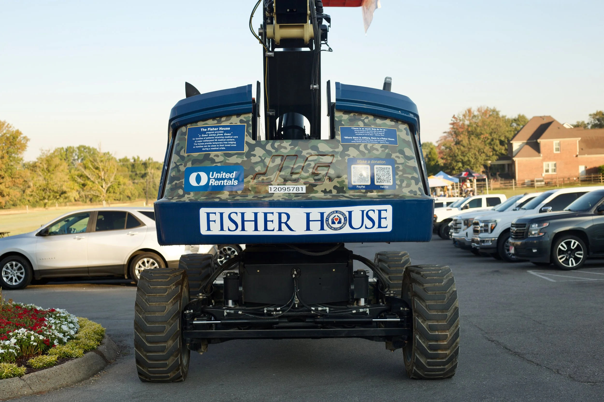 The image shows the front view of a large outdoor vehicle or piece of equipment, possibly a lawn mower or construction machinery, with the words 'Fisher House' and 'United Rentals' on it. The background includes parked cars, a building, and trees.
