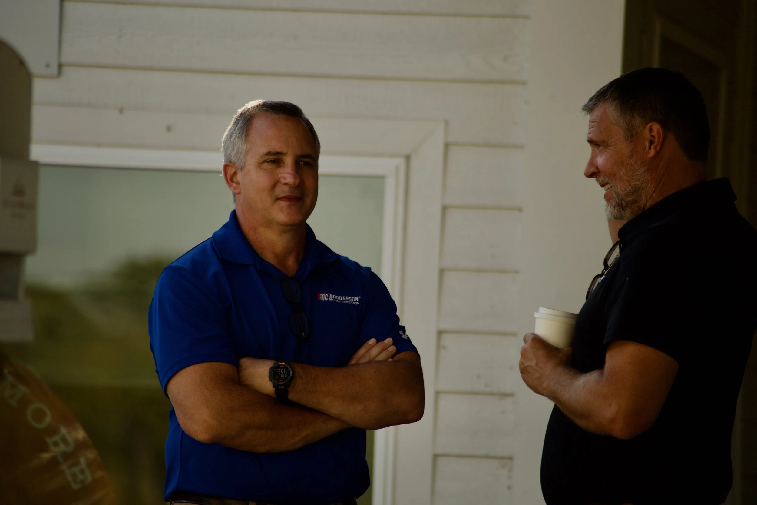Two middle-aged men are standing and talking outdoors. The man on the left is wearing a blue shirt and has his arms crossed. The man on the right, with a beard and gray hair, is holding a coffee cup and smiling while listening. The background shows a