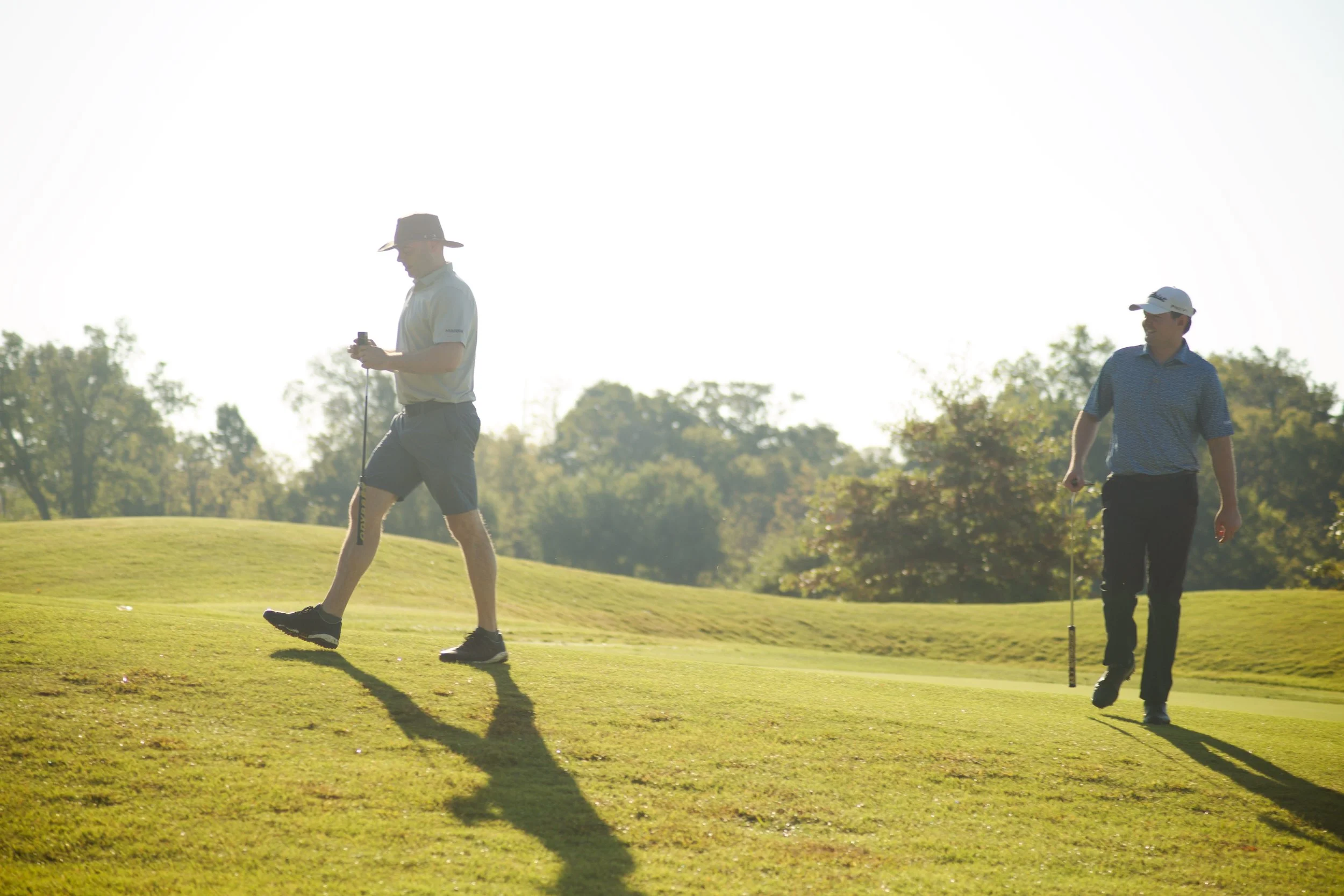 Two men on a golf course, one walking with a golf club, the other looking at his phone, during daytime with trees in the background.