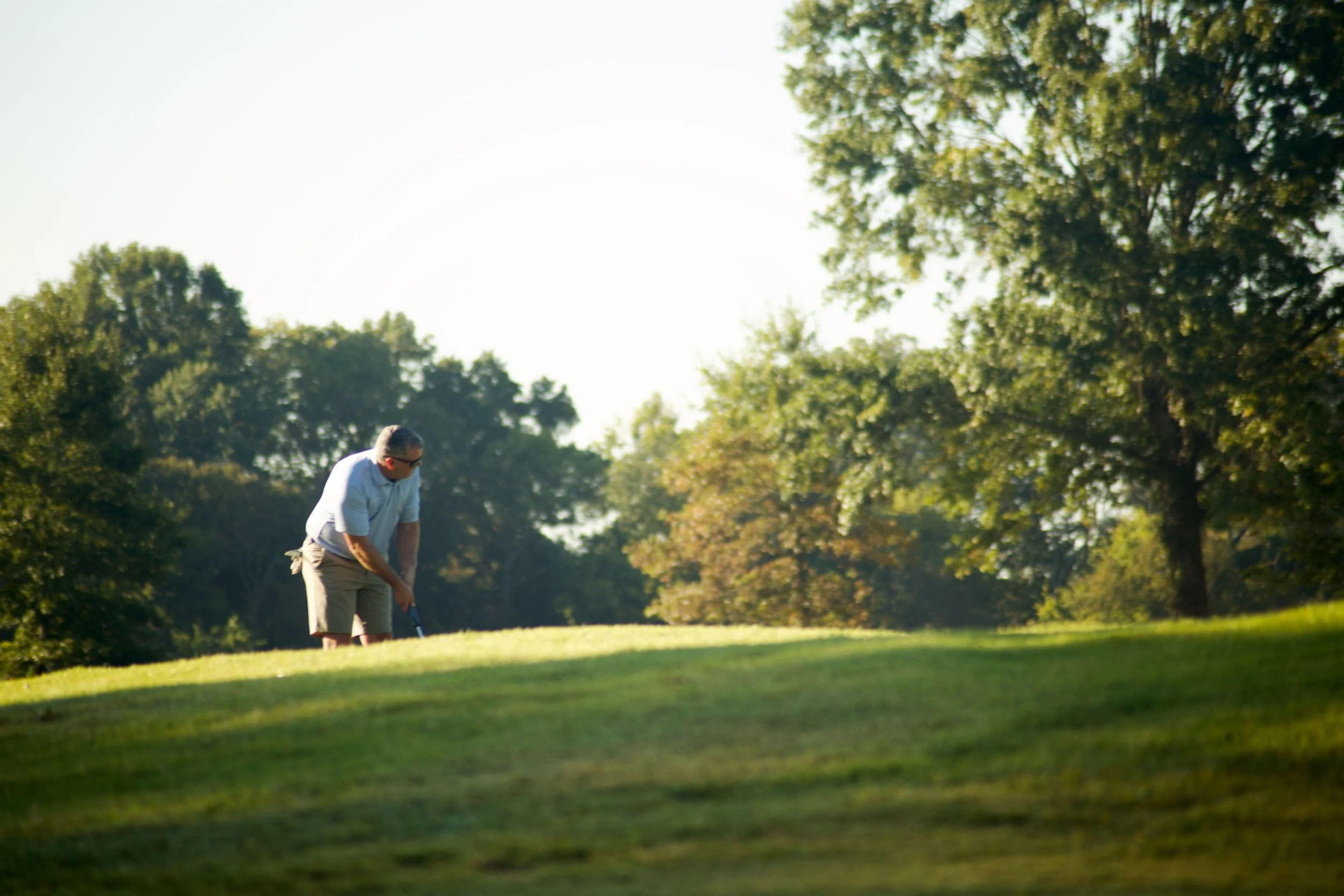 A man in a white shirt and khaki pants is preparing to hit a golf ball on a golf course with trees in the background during daytime.