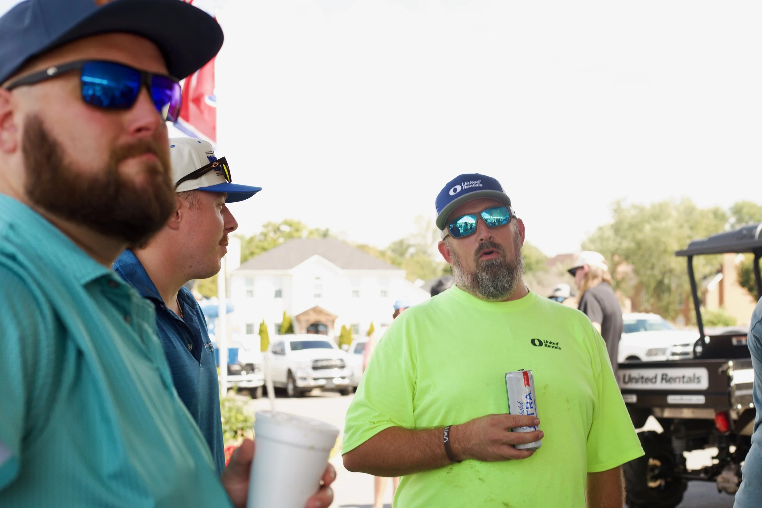A group of men talking outdoors, one in a neon yellow shirt with a 'United Rentals' logo, wearing sunglasses and a cap, holding a drink and a clipboard, with cars and trees in the background.