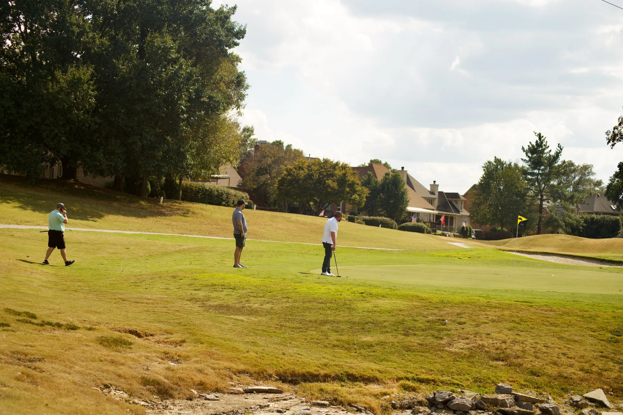 Four men playing golf on a course with houses and trees in the background under a partly cloudy sky.