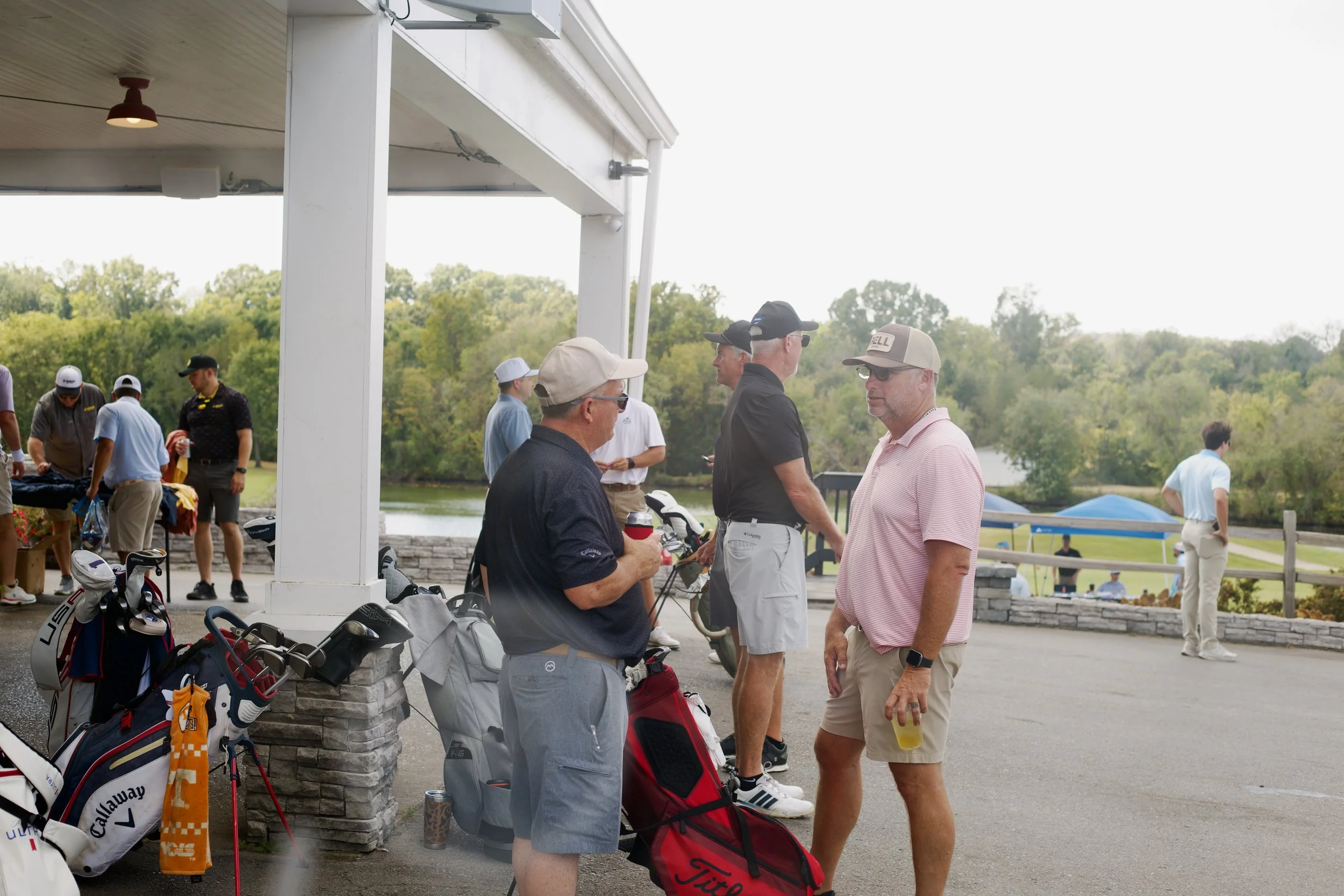 A group of men gathered outdoors near a golf course, some holding drinks and golf equipment, with trees and a lake in the background.