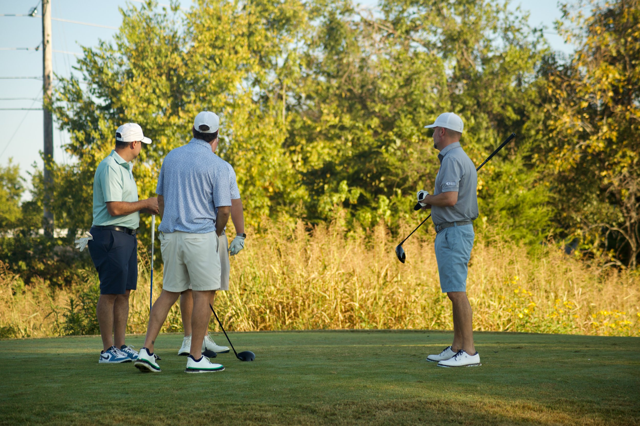 Four men standing on a golf course, discussing while holding golf clubs, with trees in the background.