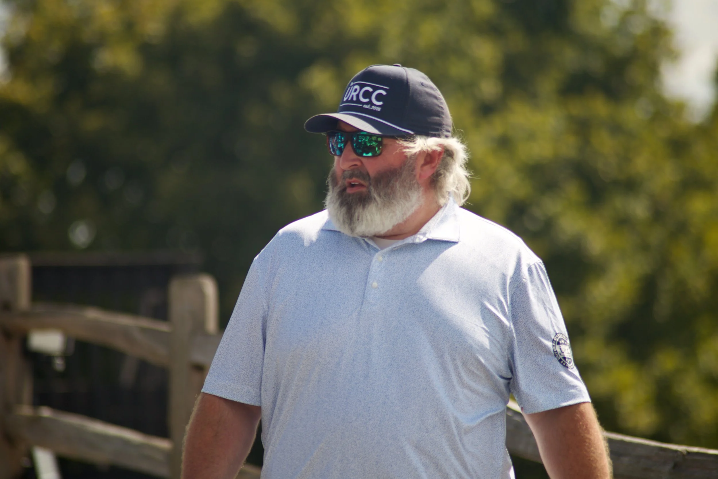 A man with white hair and beard is wearing reflective sunglasses, a navy baseball cap with 'URCC' written on it, and a light blue polo shirt with a logo on the sleeve. He is outdoors with green trees in the background.