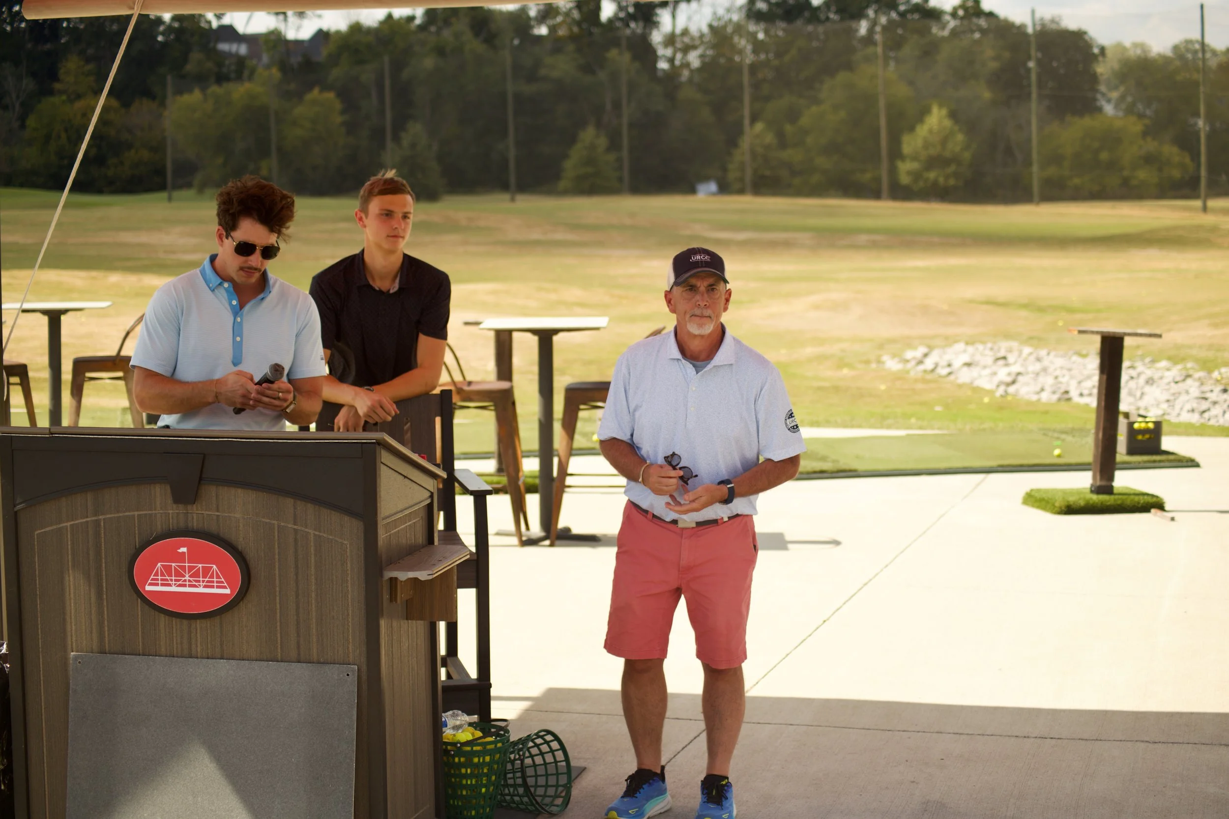 Three men standing outdoors near a wooden structure with a red logo, at a sports or recreational event, with a grassy field and trees in the background.