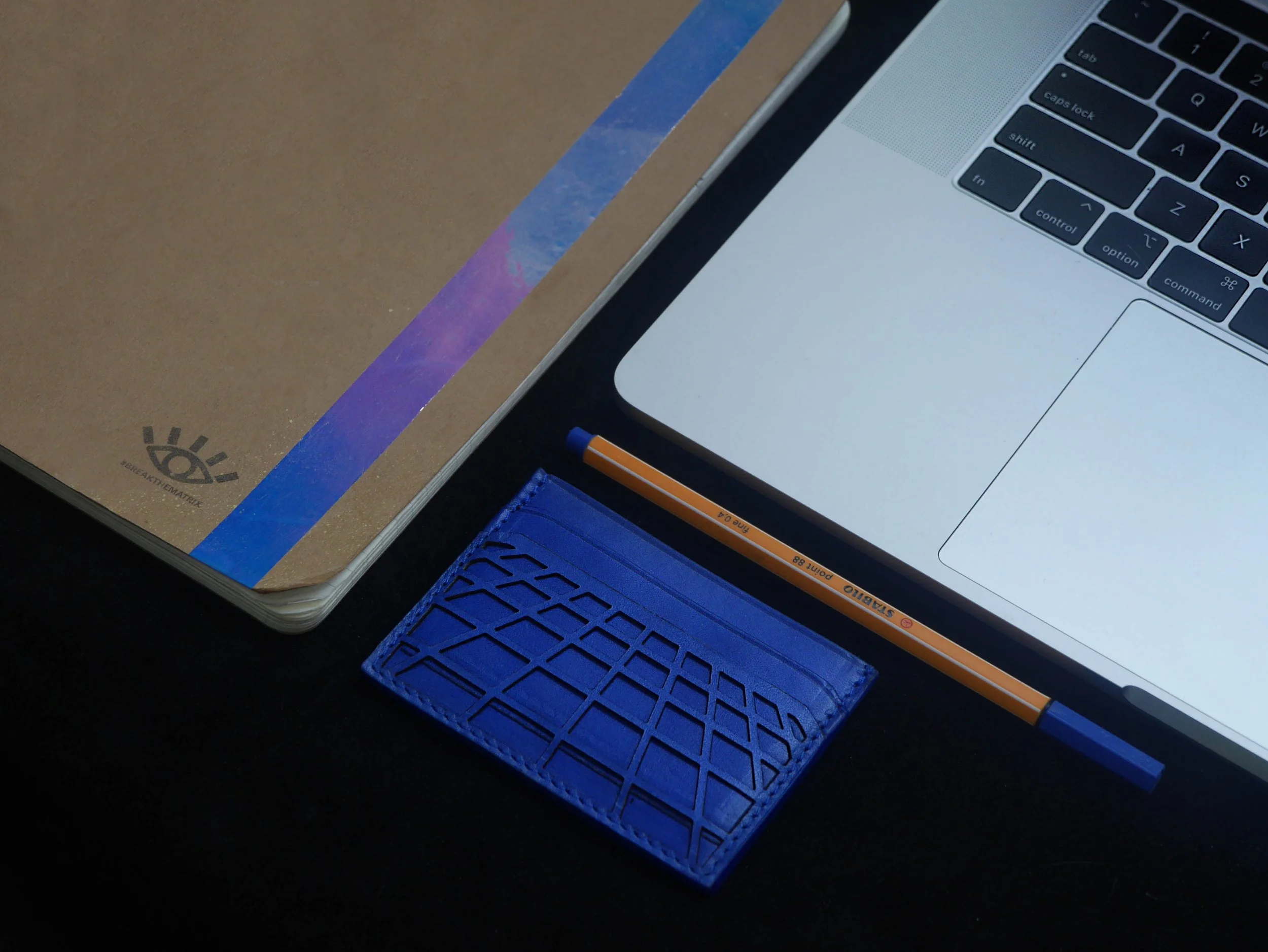 Laptop next to a brown notebook, blue cardholder, and orange pencil on a dark surface.