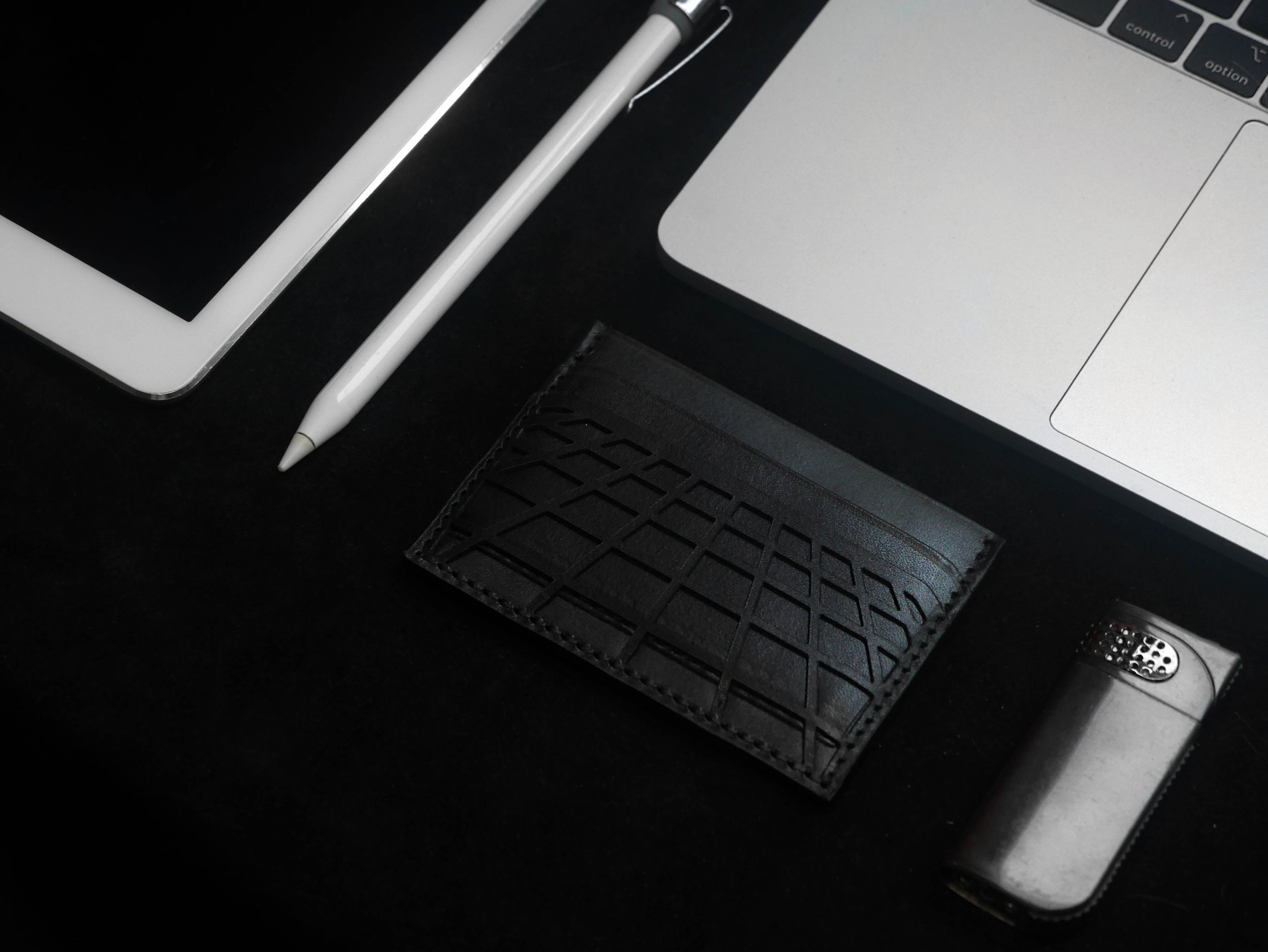 Overhead view of a digital tablet, stylus, laptop, black cardholder, and lighter on a dark surface.