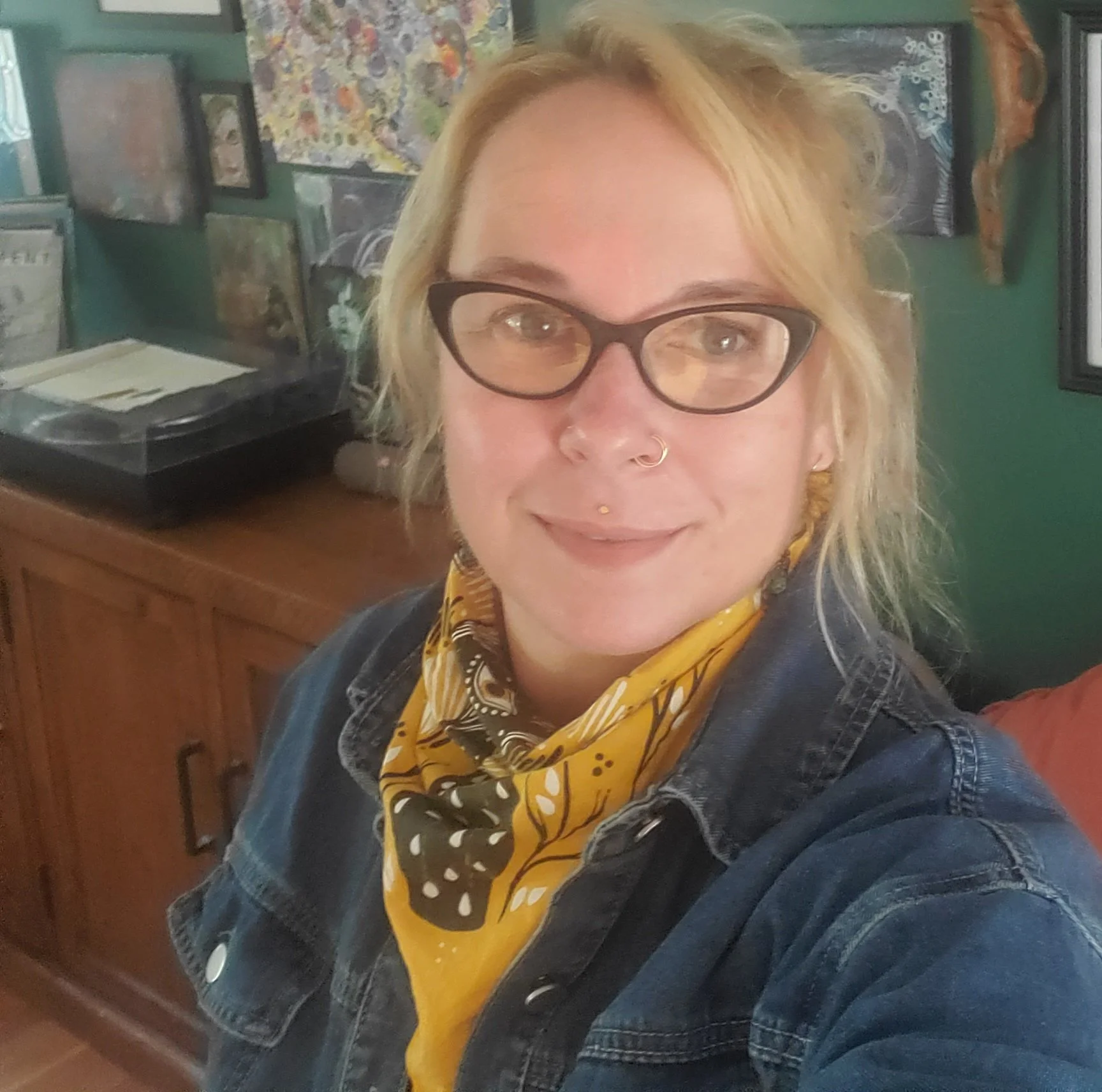 A woman with blonde hair wearing glasses, a yellow bandana, and a denim jacket, smiling at the camera indoors with artwork on the green wall behind her.