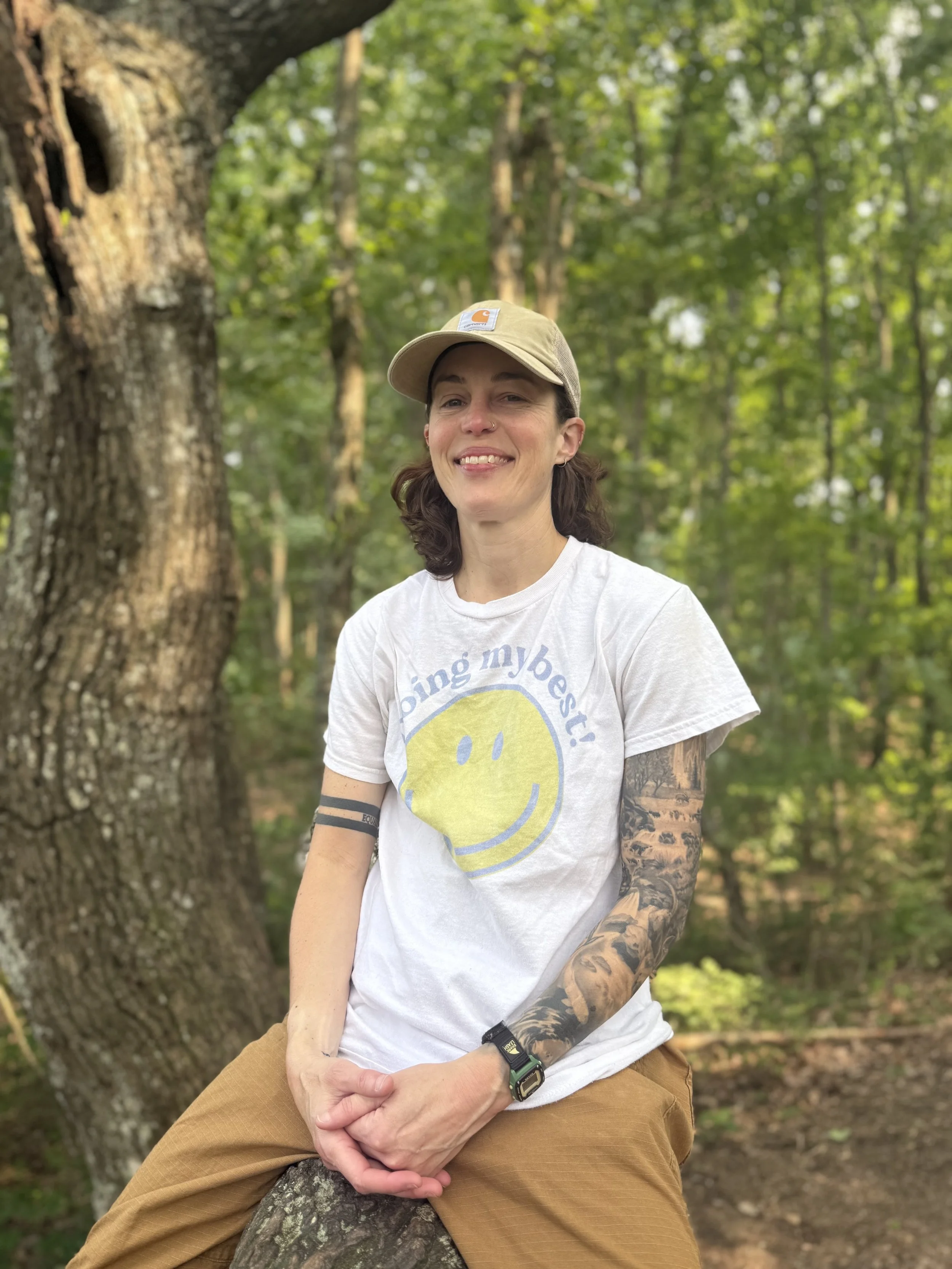 A woman sitting outdoors on a fallen tree trunk, smiling at the camera, in a forest with green trees in the background, wearing a white t-shirt with a yellow smiley face and the text 'Doing my best,' a beige cap, tan pants, and sporting tattoos and a smartwatch.
