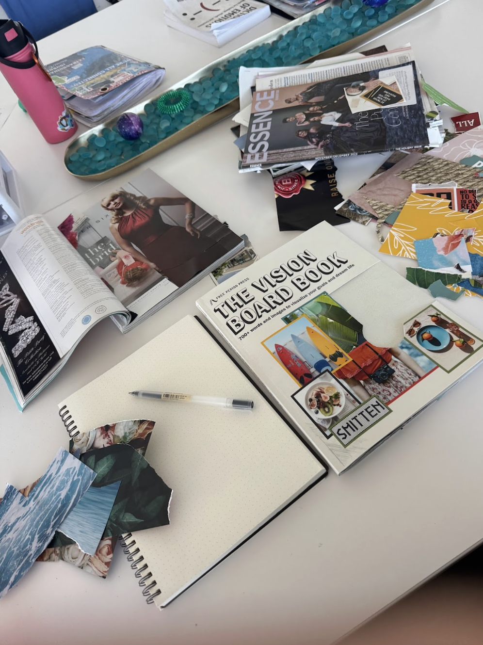 Creativity taking place in an art workshop led by Dr. Jenna Freeman - white table with magazines, a spiral notebook, a pen, a pink water bottle with a black lid, decorative stones, a green bracelet, printed photos, and cut-out images and papers.