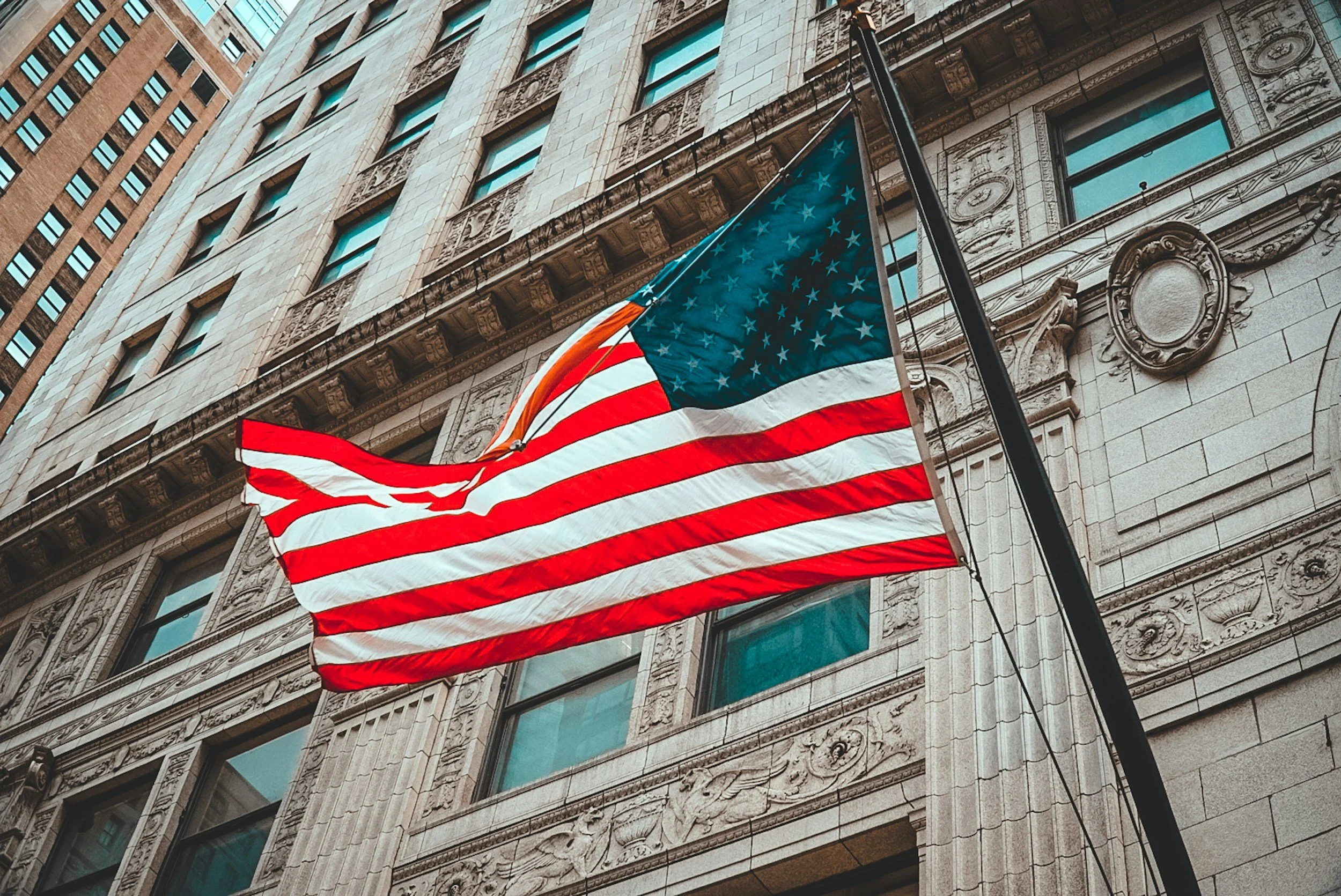 United States flag waving in front of a historic stone building with ornate architectural details and multiple windows.