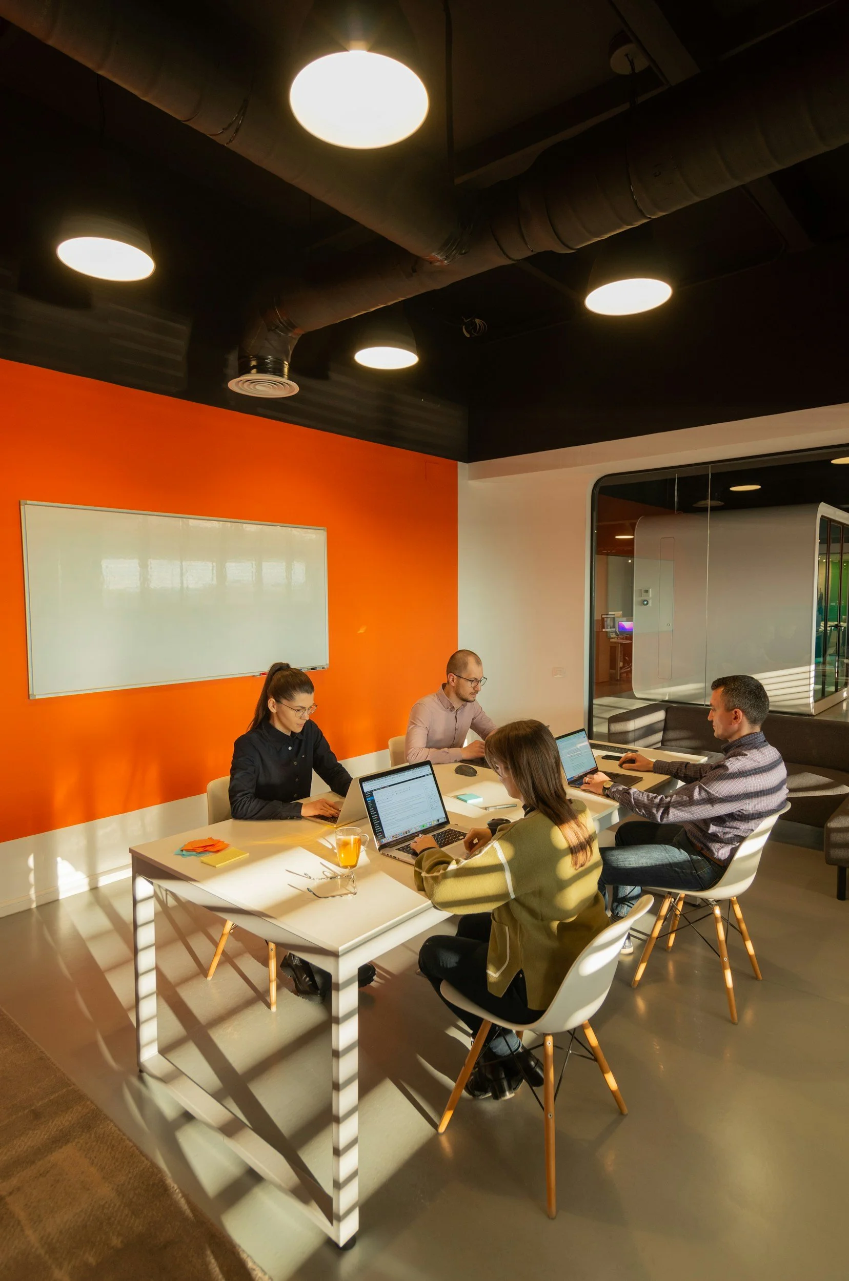 Four people working on laptops at a business meeting in a modern office with an orange accent wall and overhead lighting.