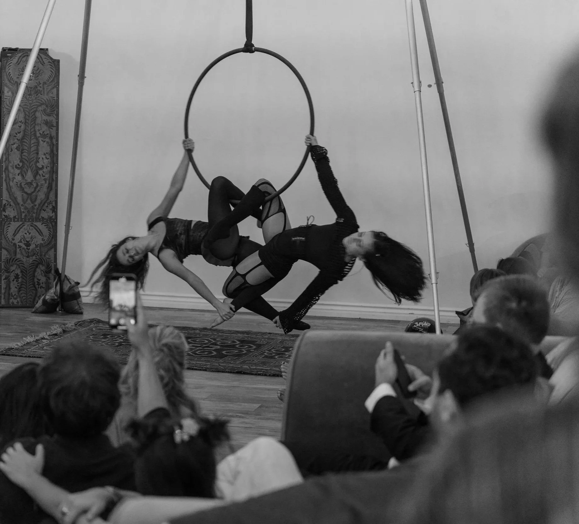 Two women perform a circus aerial act on a hoop, while an audience watches and takes photos.