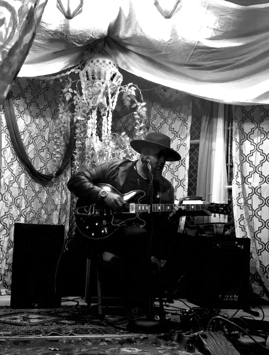 A man wearing a large hat is playing an electric guitar and singing into a microphone in a decorated indoor setting with patterned curtains and drapery.