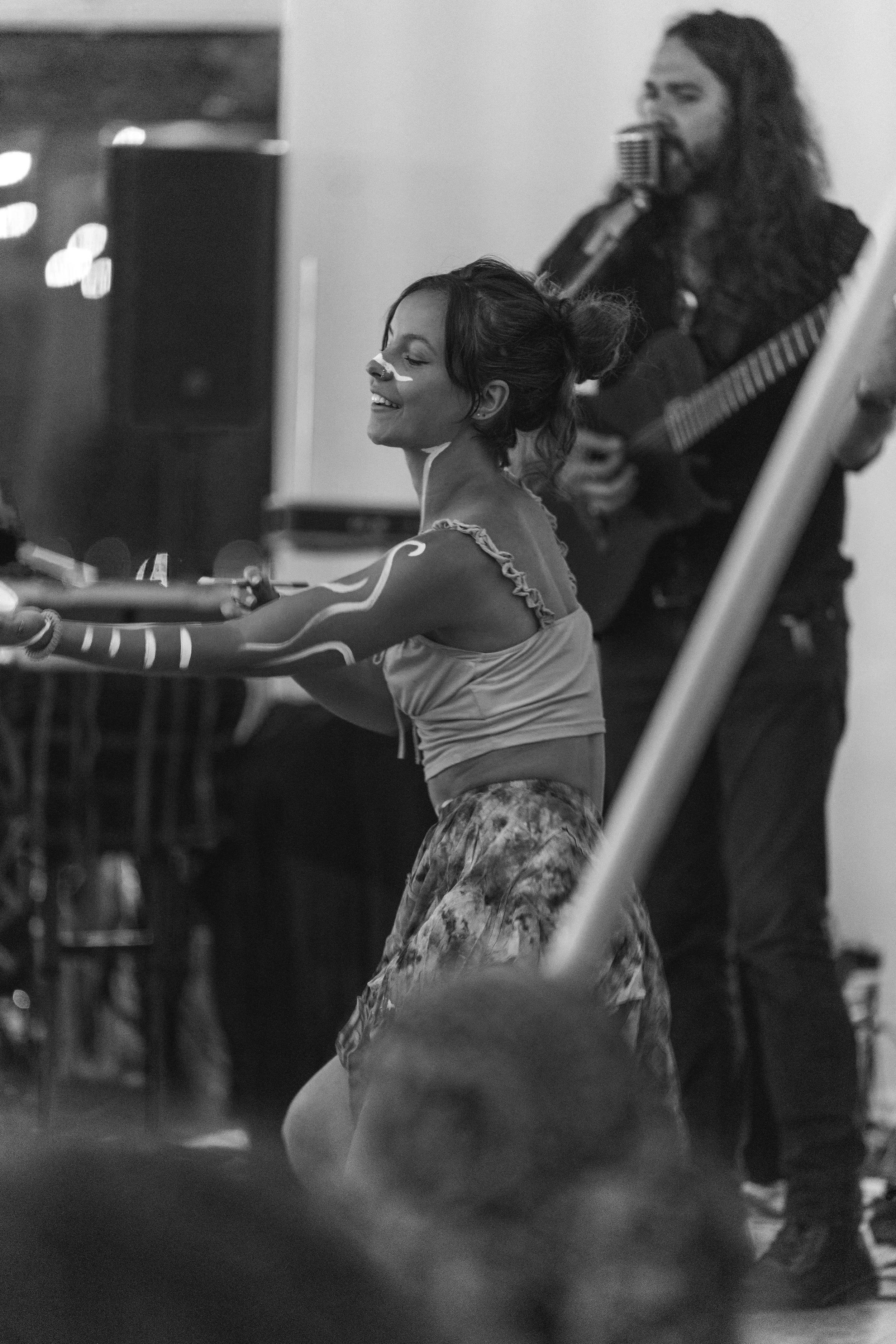 A woman smiling and dancing with body paint on her arms, while a man plays guitar and sings in the background, in a black and white indoor setting.