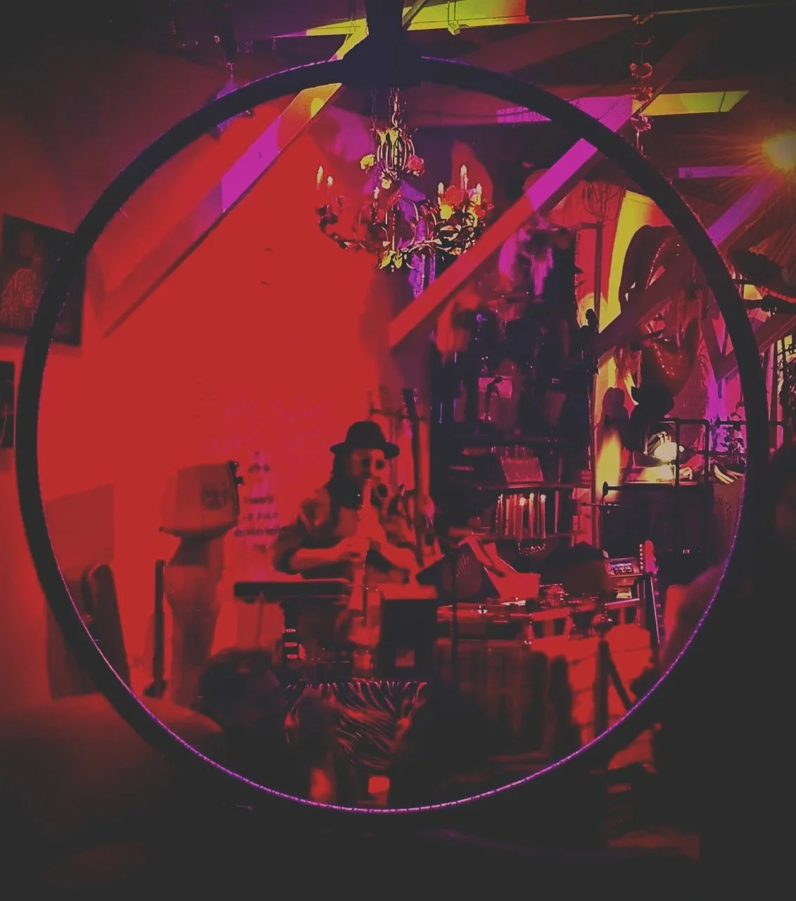 Reflective round mirror showing a dimly lit room with a chandelier, people, and various artwork and furniture, with red and purple neon lighting.