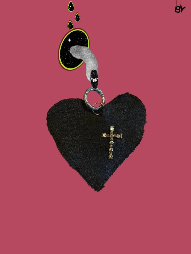 A black heart-shaped plush with a cross embellishment hanging from a metal ring, set against a pink background with a sticker of a black and white cartoon snake and black droplets above.