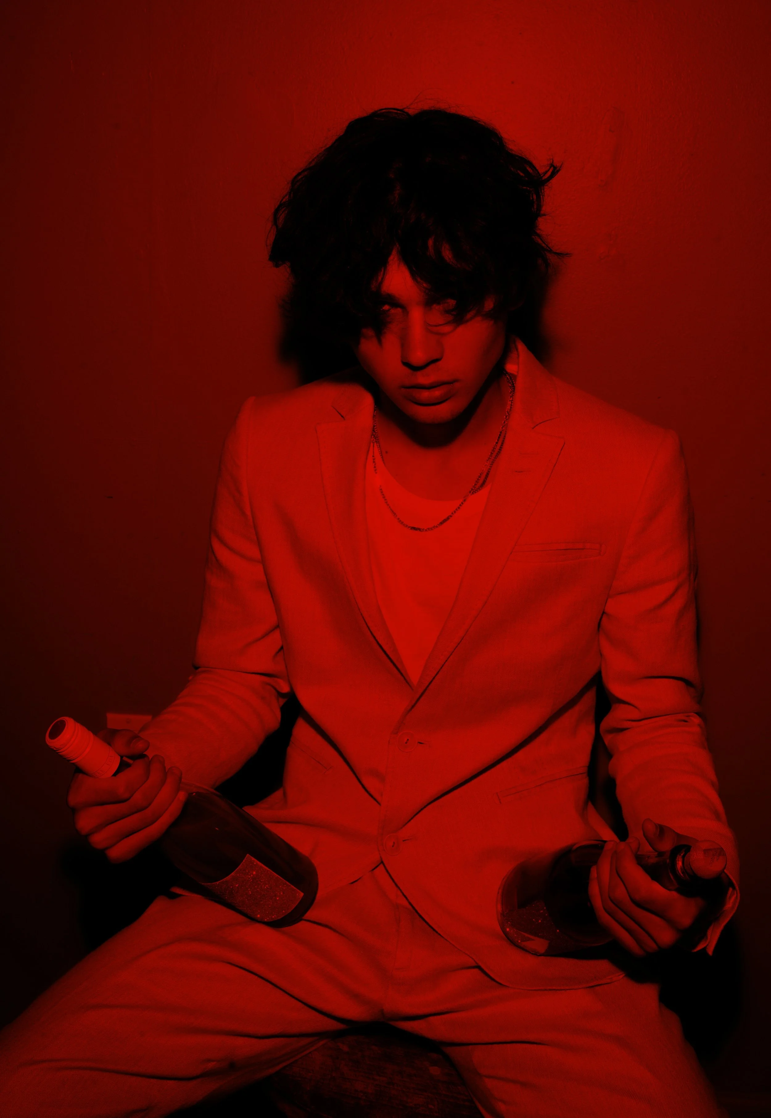 A person with dark, messy hair wearing a light-colored suit and a layered necklace, sitting against a red background, holding a bottle and a glass.