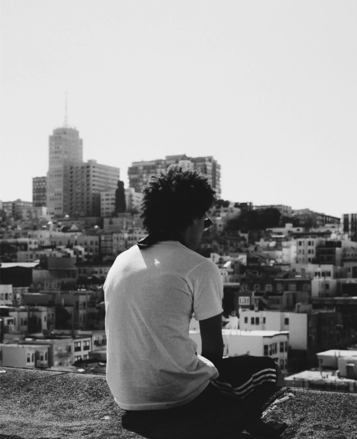 A person with curly hair and sunglasses sitting on a rooftop ledge overlooking a cityscape with tall buildings.