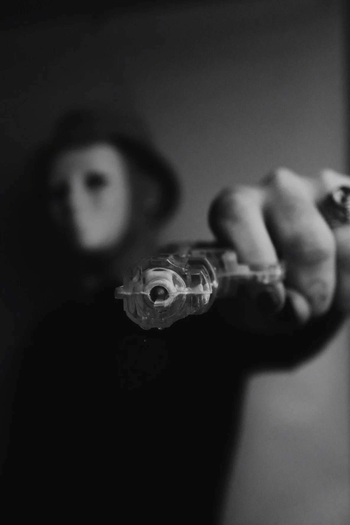 A person holding a gun pointed towards the camera, with a blurry face in the background in black and white.