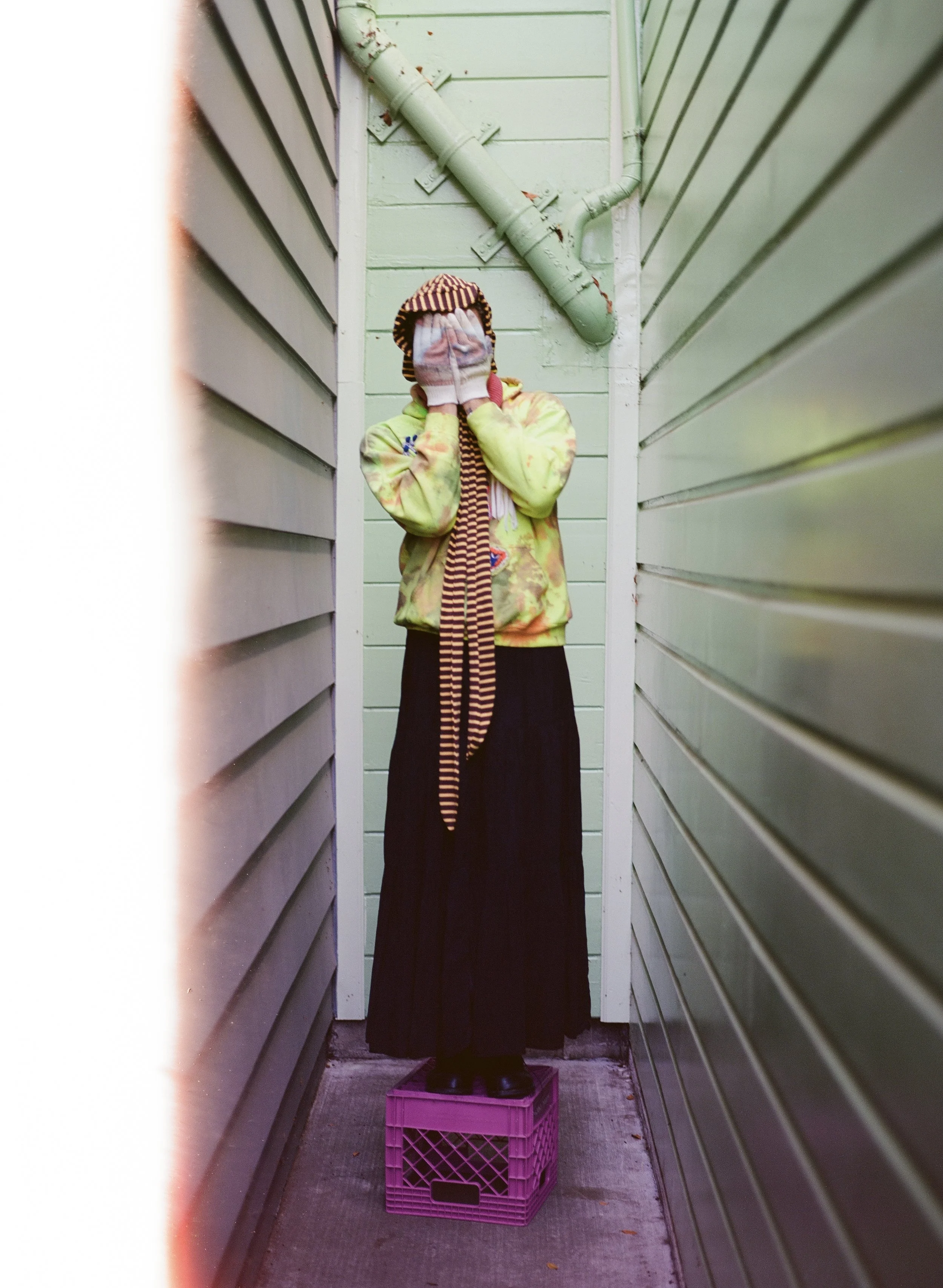Person standing on a pink crate in a narrow alleyway with siding on both sides and a green drainpipe on the back wall, covering their face with their hands.