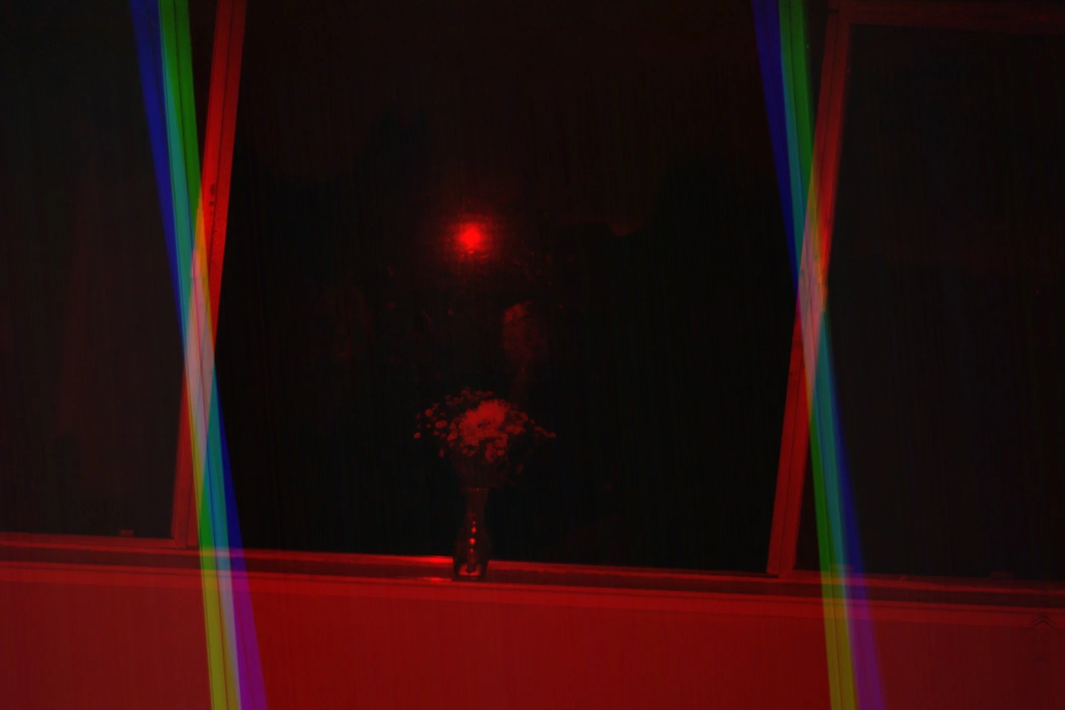 A window with colorful neon light reflections in red, green, blue, and purple, and a vase with flowers on the windowsill, illuminated by a red light in a dark room at night.