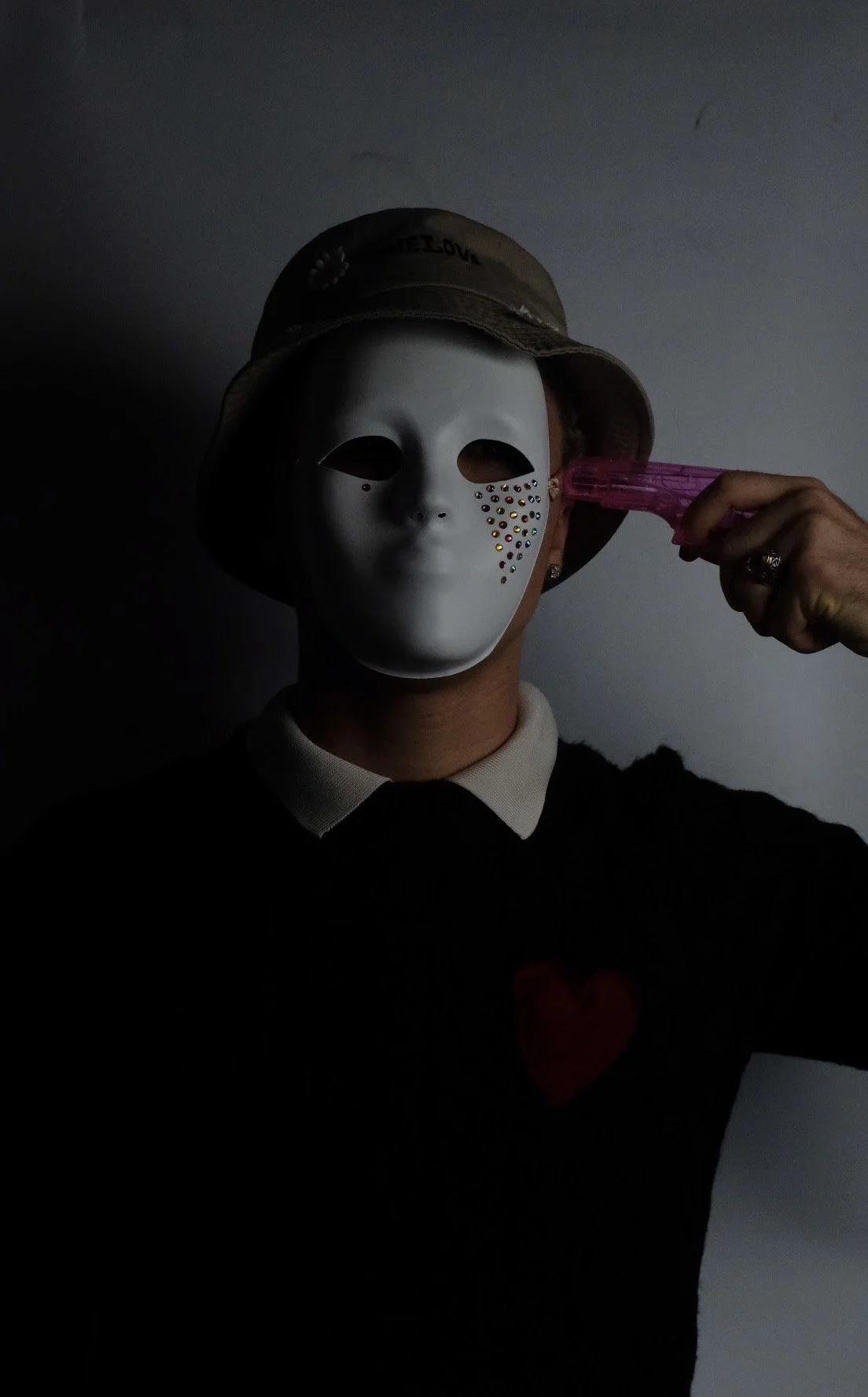 Person wearing a mask with rhinestones and a beige hat, holding a pink gun to their head.