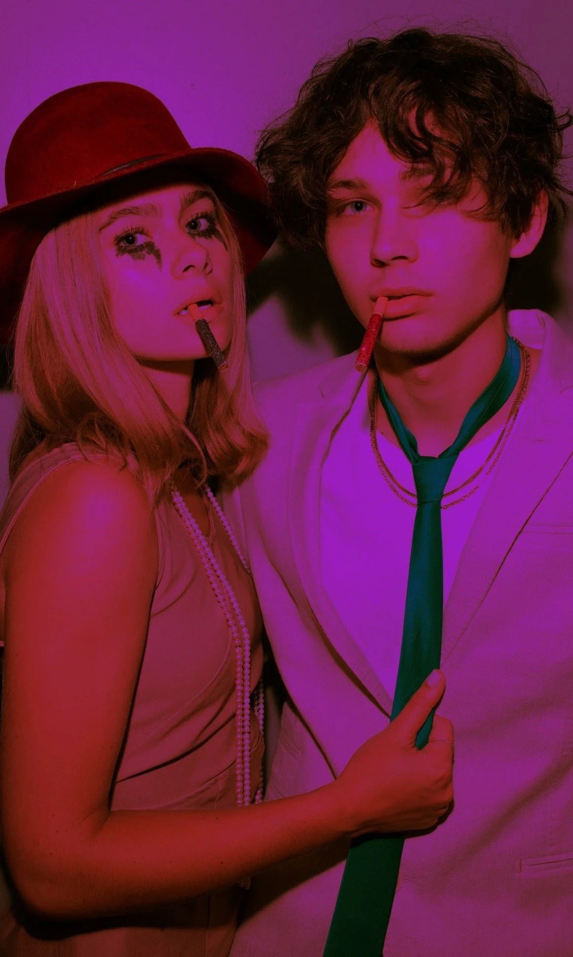 A young woman and man dressed in vintage 1960s style, with the woman wearing a brown hat and pearl necklace and the man in a suit with a tie, both holding candy sticks in their mouths, in a retro-themed setting with pink and purple lighting.