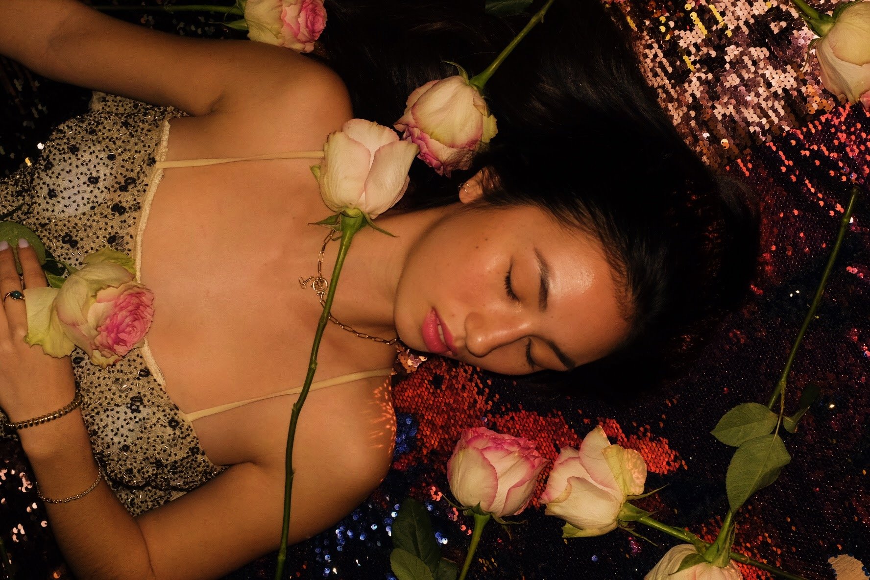 A woman with long dark hair lying on a colorful, sequin-covered surface, surrounded by pink and white roses, with her eyes closed and a slight smile.