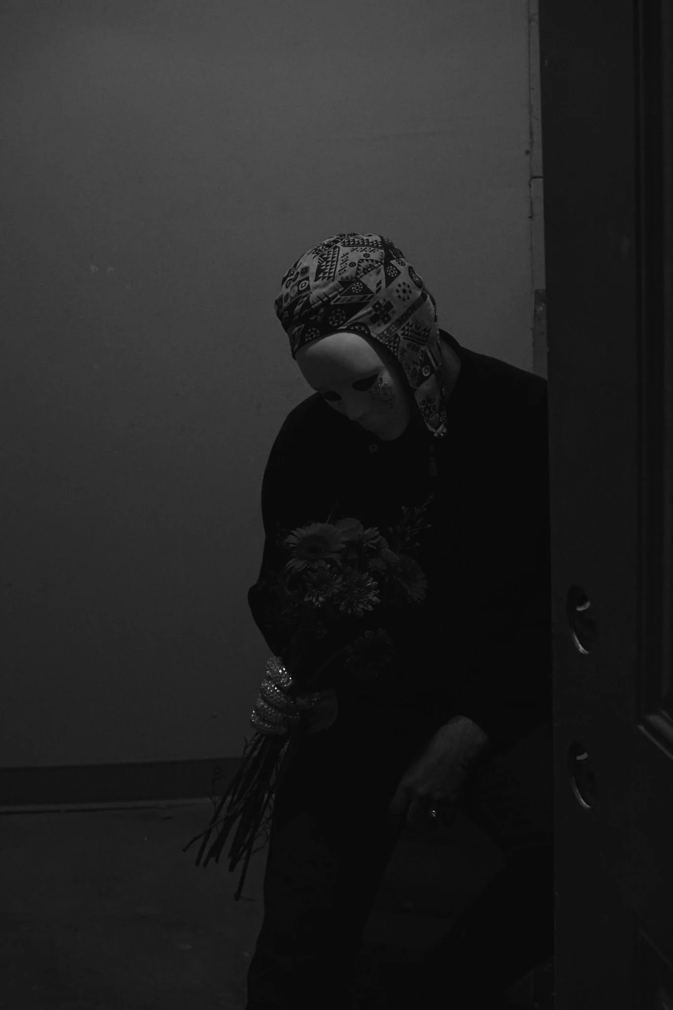 Person wearing a mask and headscarf holding a bouquet of flowers, dark indoor setting, black and white photo.