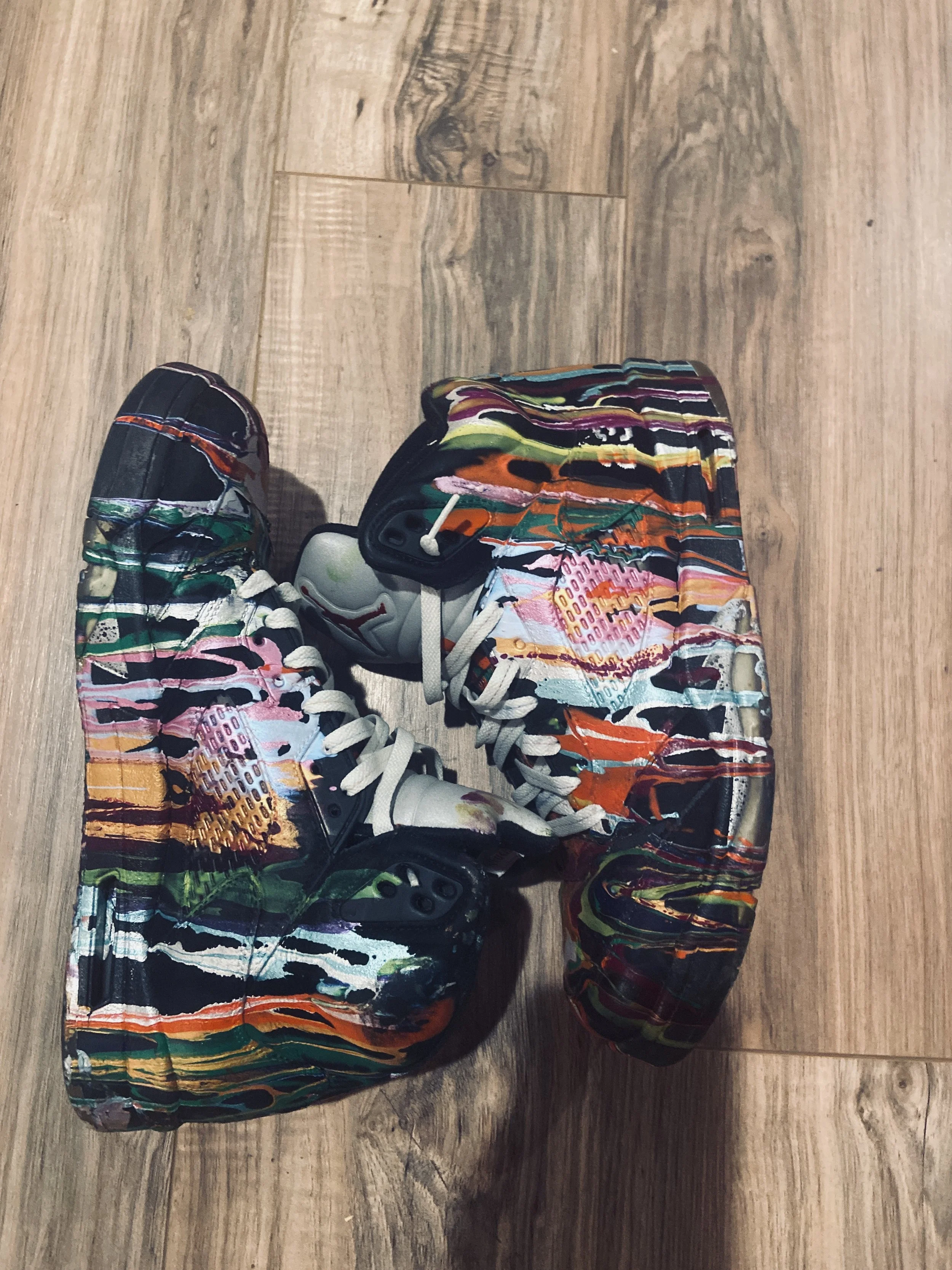 A pair of sneakers with a colorful, abstract, painted design on the upper and soles, placed on a wooden floor.