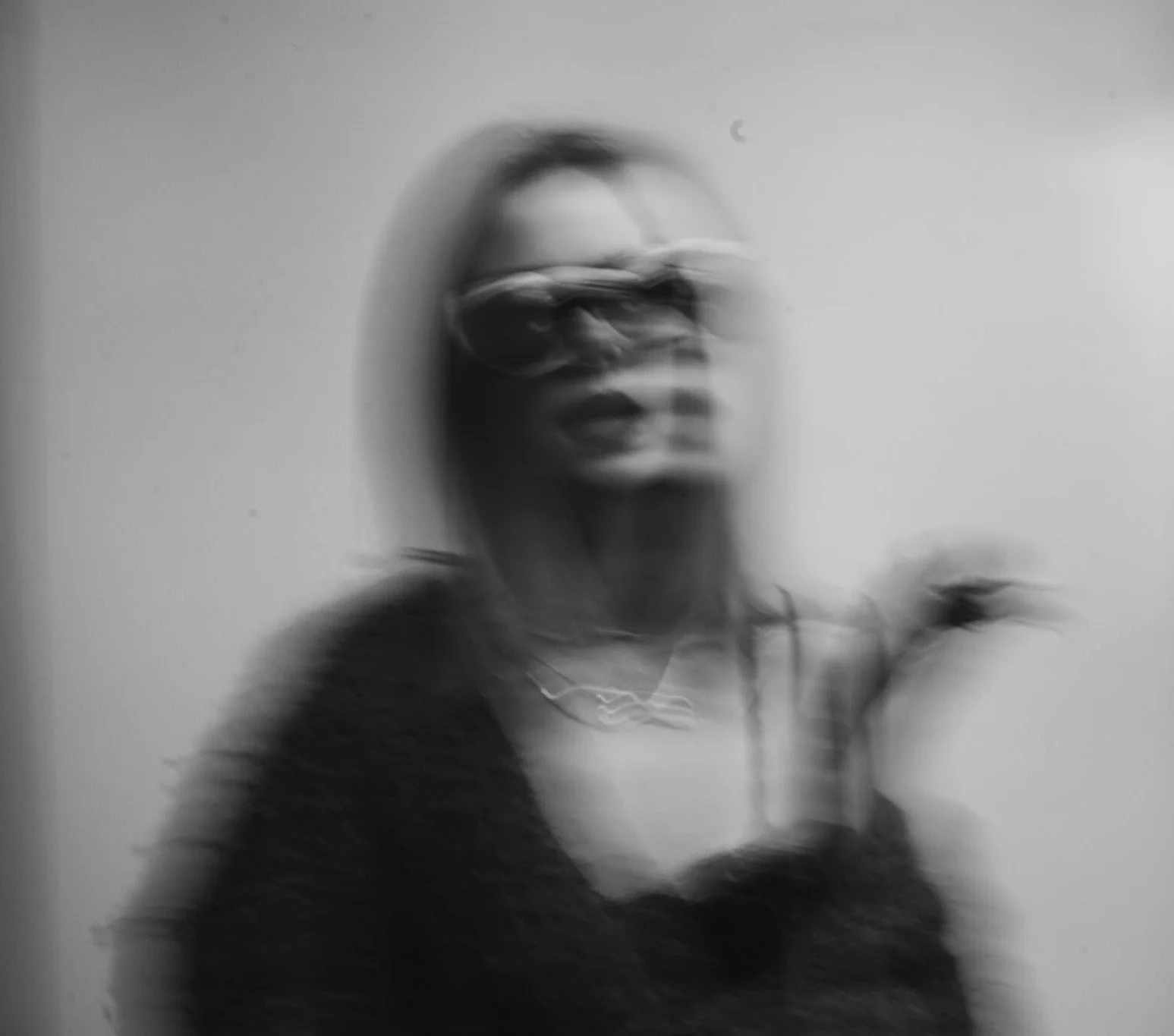 Blurred black-and-white photo of a woman with sunglasses and jewelry, standing against a plain wall, holding something in her hand.