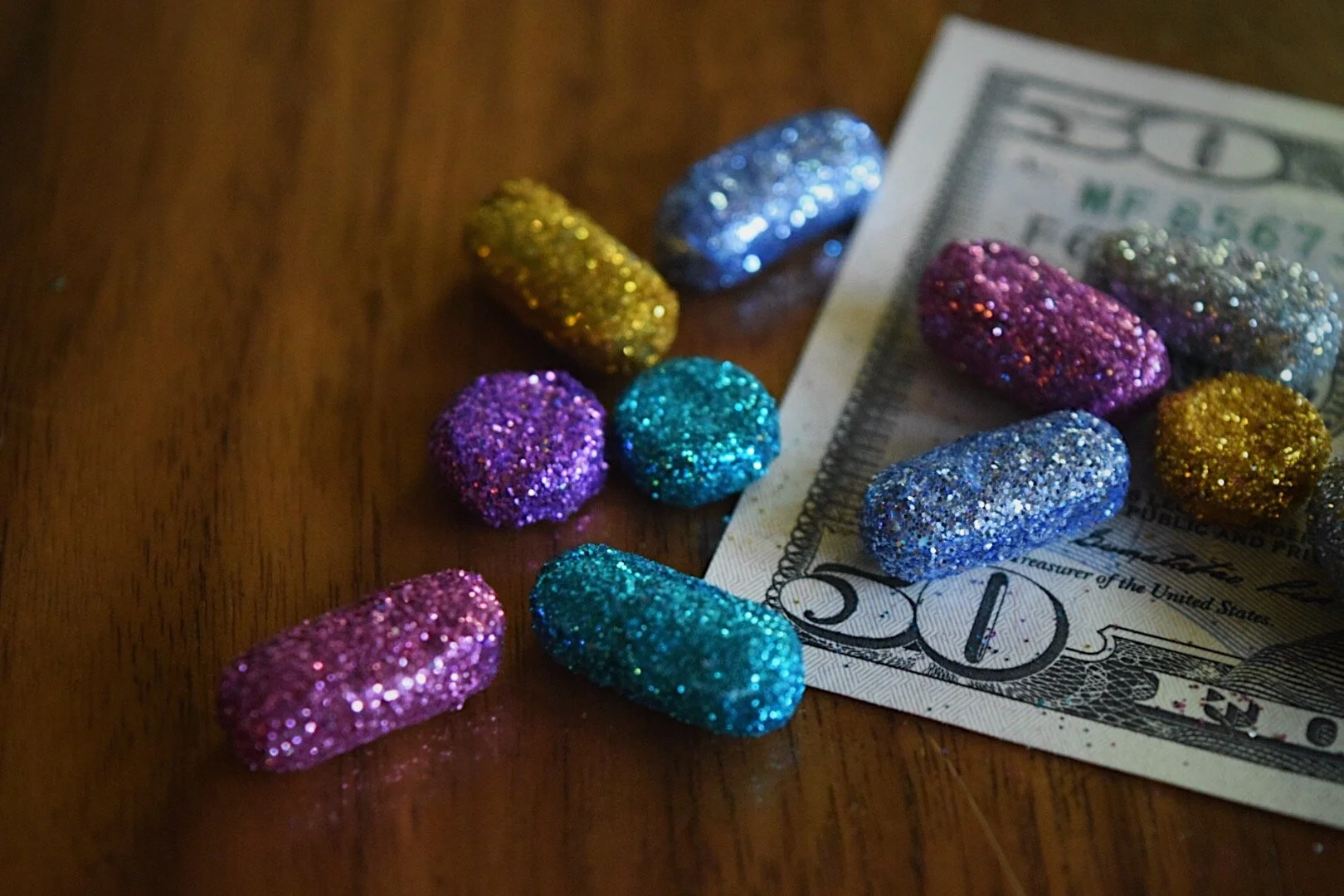 Colorful glitter-coated pills scattered on a wooden surface with a fifty-dollar bill underneath some of them.