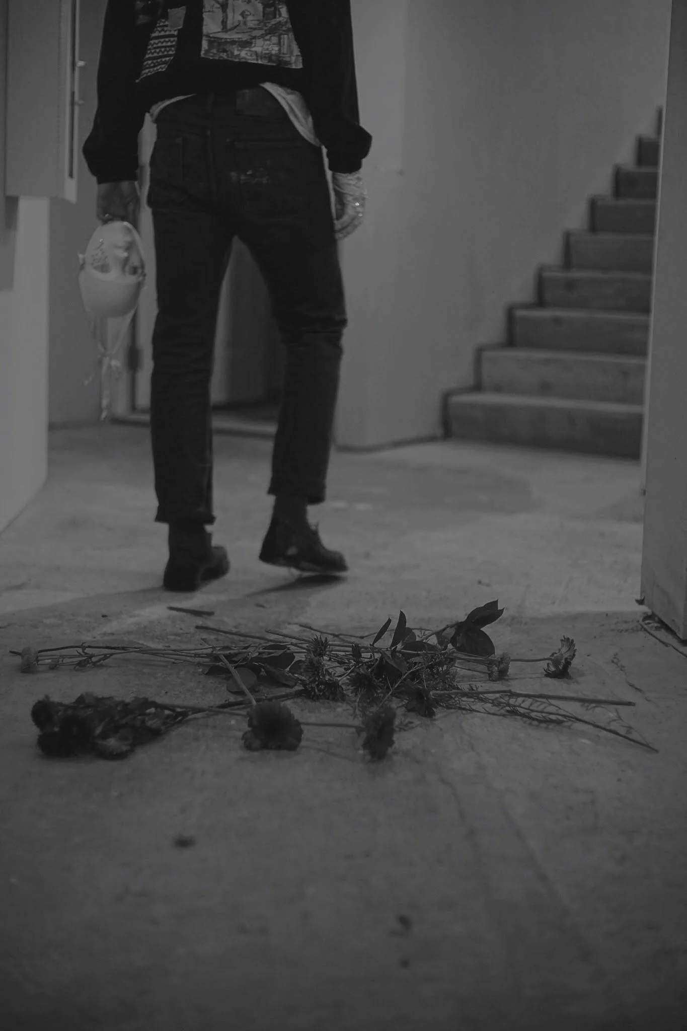 Person walking away holding a mask in their right hand, seen from behind, with wilted flowers on the floor.