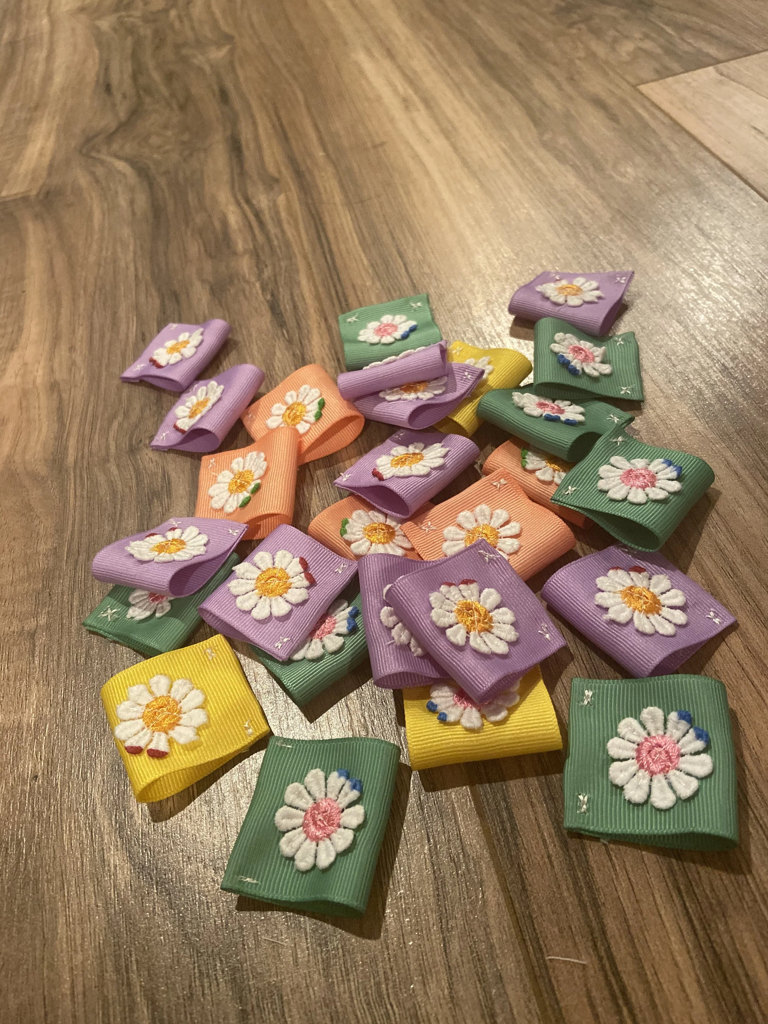 Colorful fabric ribbon bows with a daisy flower embroidered on each, scattered on a wooden floor.