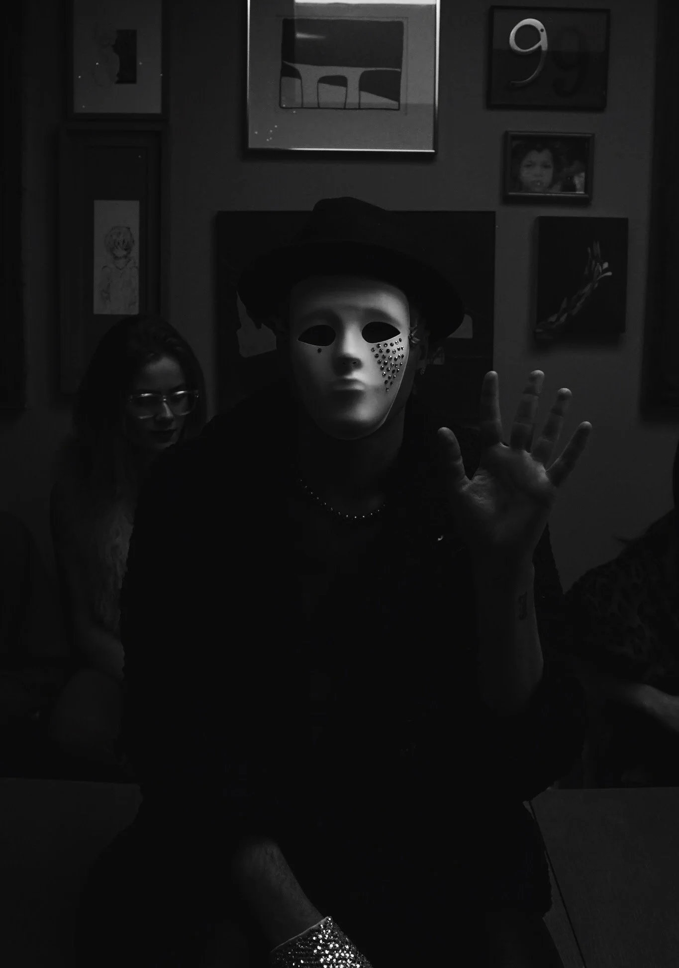 Person wearing a white mask with black accents and rhinestones, wearing a black hat, sitting and reaching out with their hand, in a room with framed artwork on the wall behind.