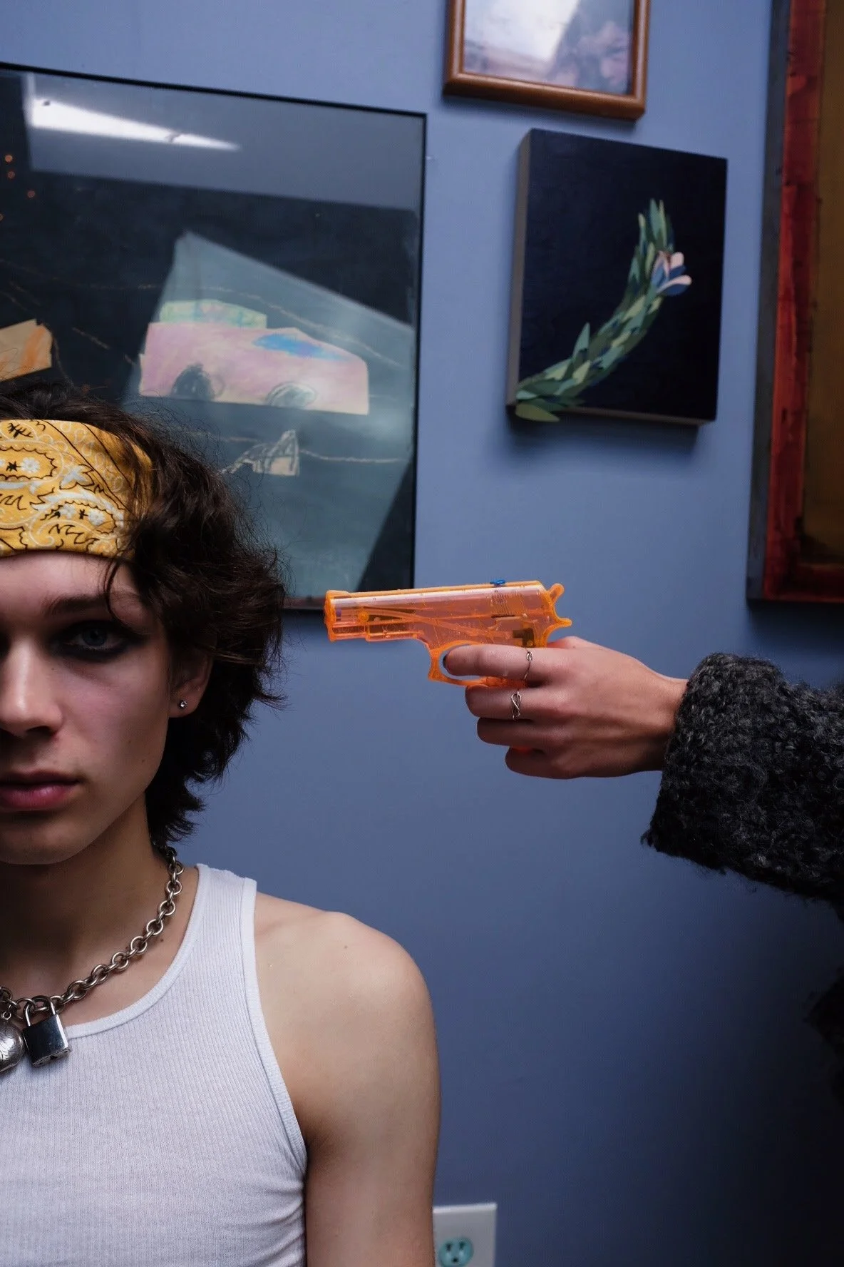 A person with a yellow bandana and white tank top has a serious expression while a hand holding an orange toy gun points it at their head. The background features a blue wall with several framed artworks.