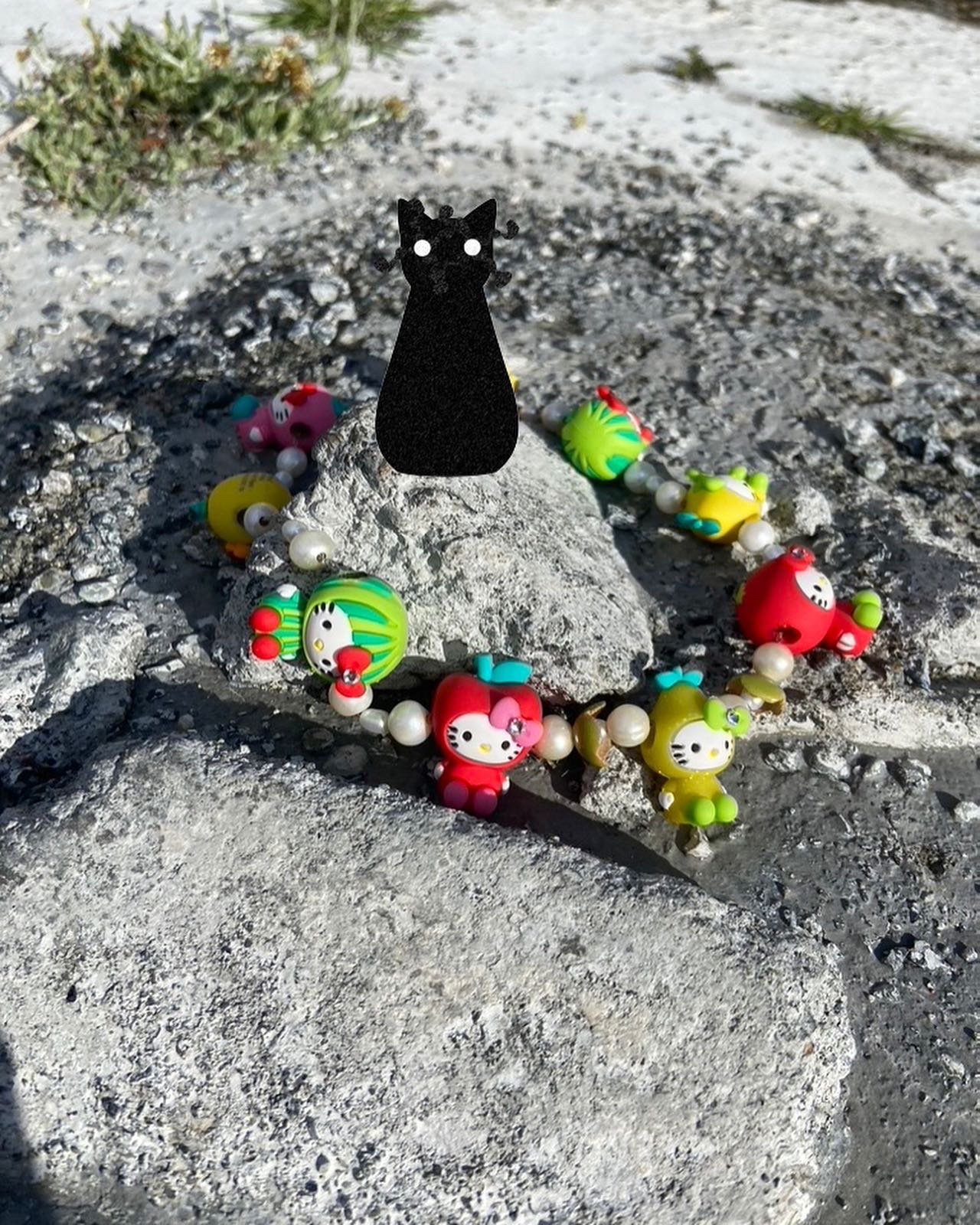 A black cat-shaped figure with cut-out eyes placed on a large rock, surrounded by a colorful beaded necklace with Hello Kitty and fruit-shaped character beads, outdoors on a rocky surface.