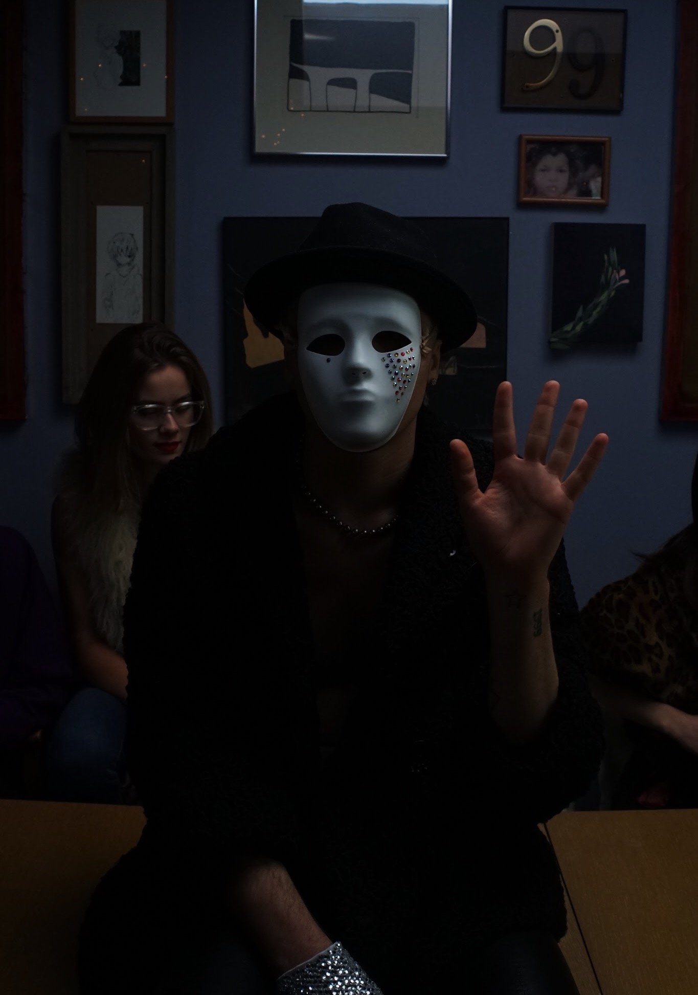 Person wearing a white mask with rhinestones, a black hat, and black clothing, waving while seated indoors with artwork and photos on the wall behind.
