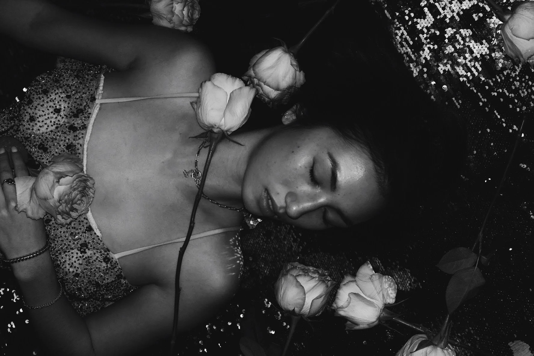 A woman with dark hair and closed eyes lying down surrounded by roses.