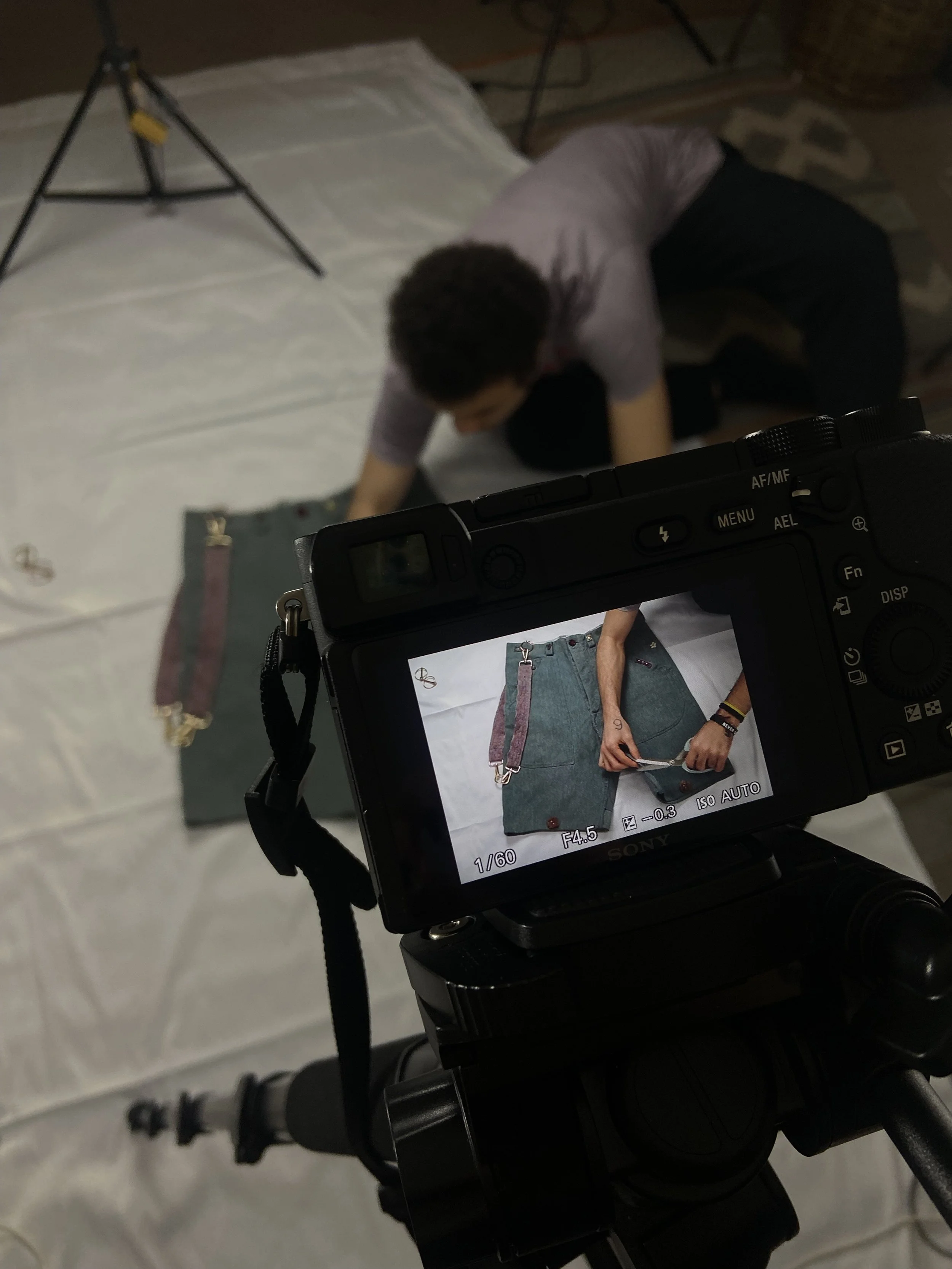 A person is kneeling on the floor, working on a photo shoot setup. The camera captures an image of a pair of denim shorts laid flat on a white surface, with a person holding a small device or object inside the shorts.