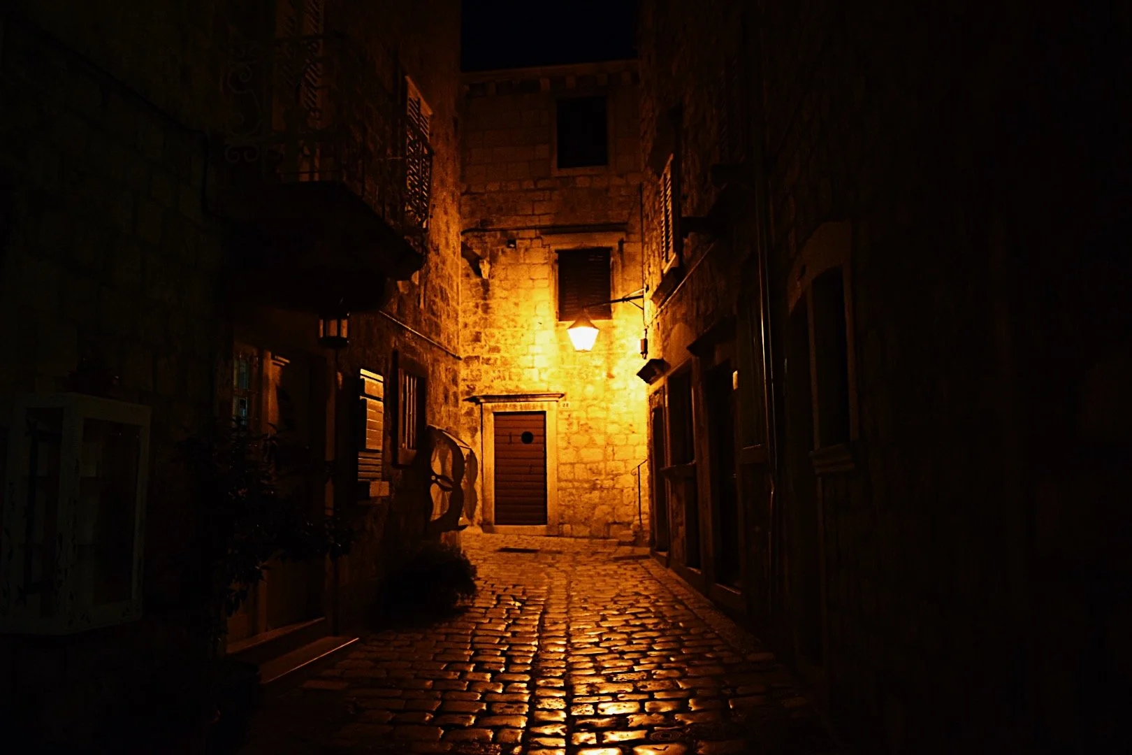 A narrow cobblestone alley at night illuminated by a single wall-mounted streetlamp, with stone and brick buildings on either side.