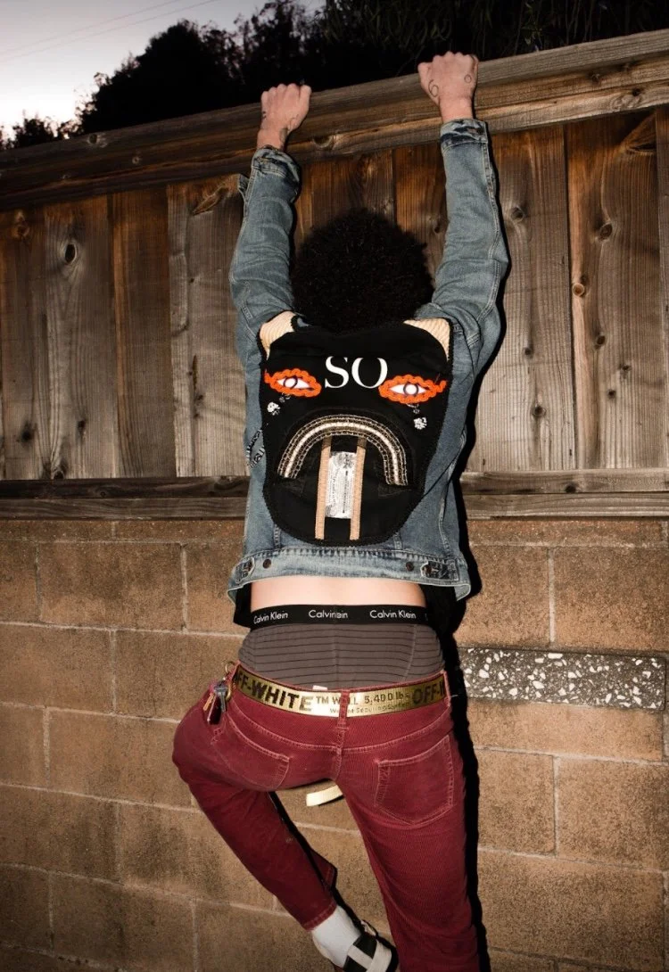 Person hanging with arms up, facing a wooden fence. Wearing a black jacket, red pants, Calvin Klein underwear waistband visible, and a backpack with eyes and rainbow design.