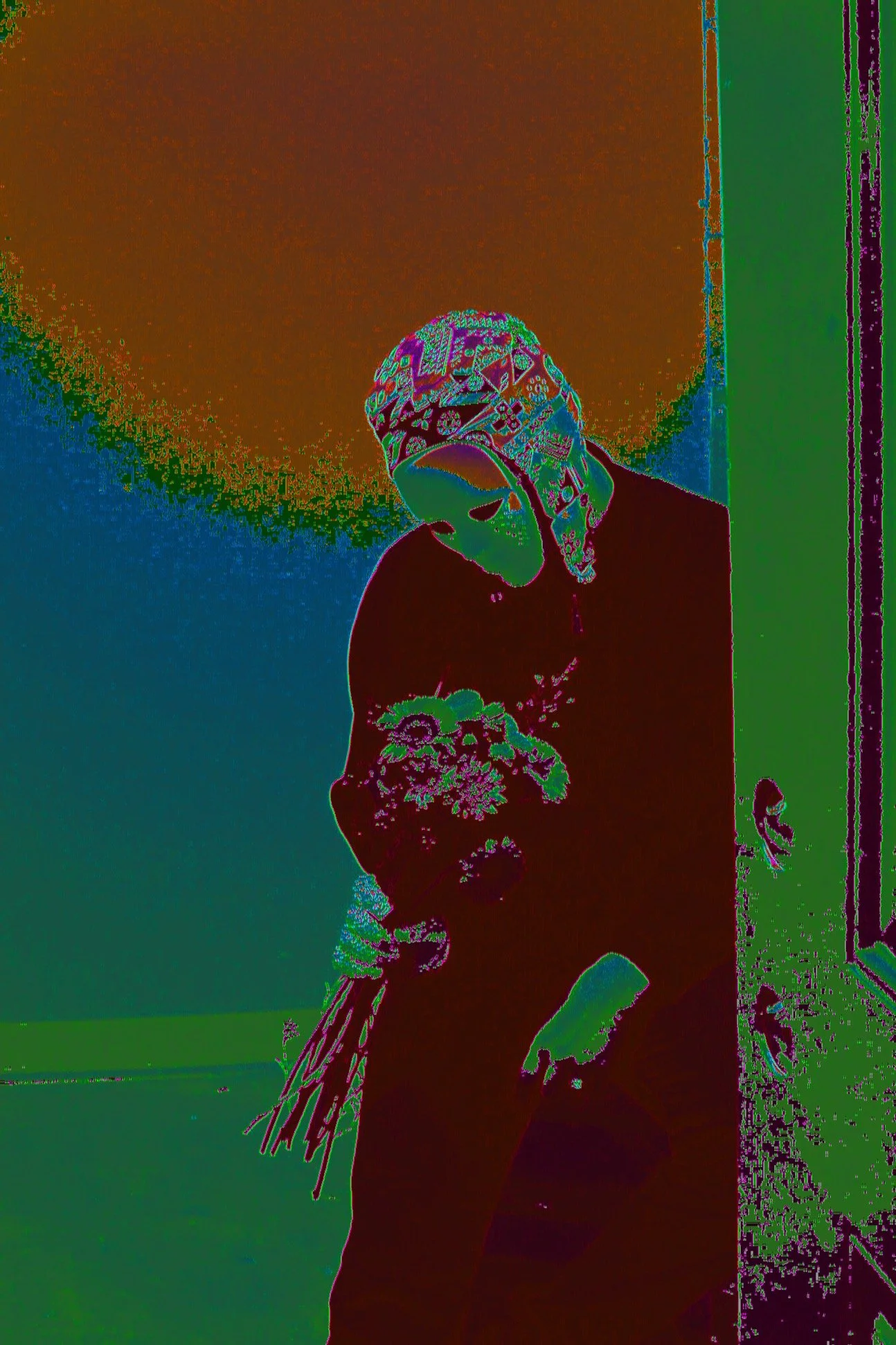 Person wearing a patterned bandana and a face mask, holding flowers, standing near a doorway.