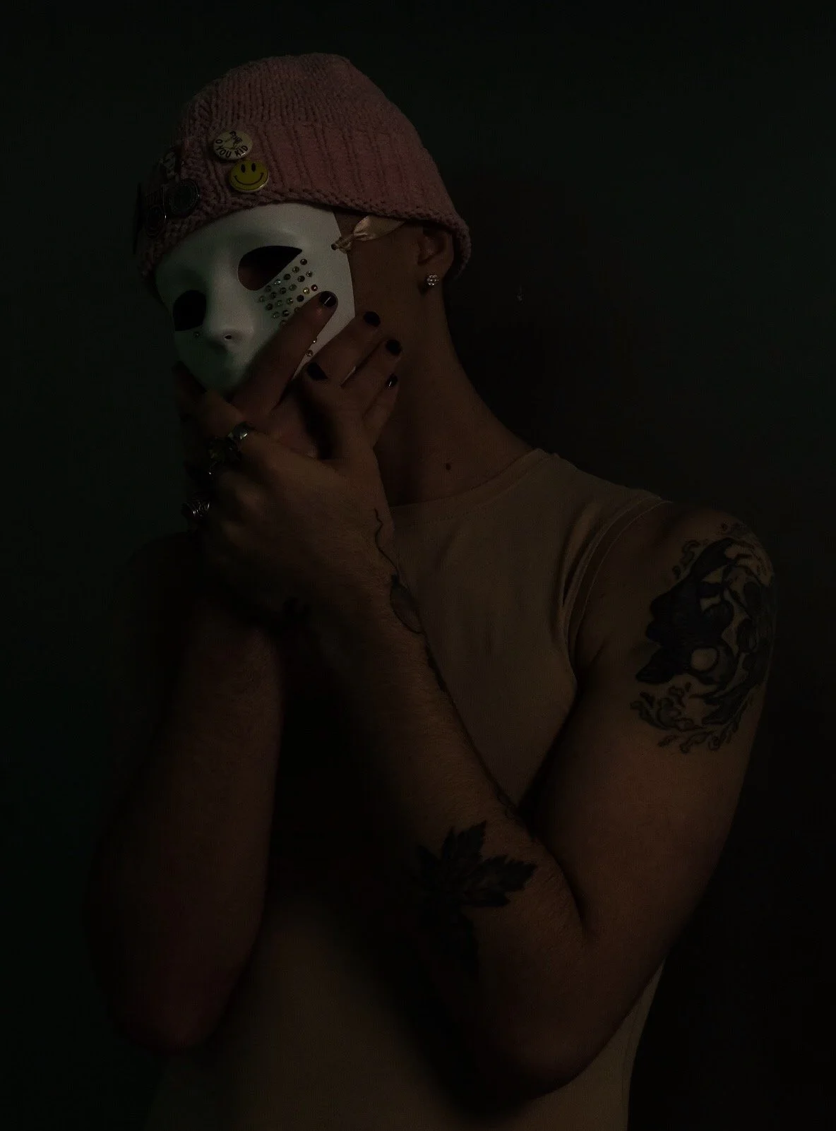Person wearing a pink knit cap, a white mask with a grid and holes, and various pins, posing with hand to face, tattoos on arms, and wearing a beige shirt, against dark background.